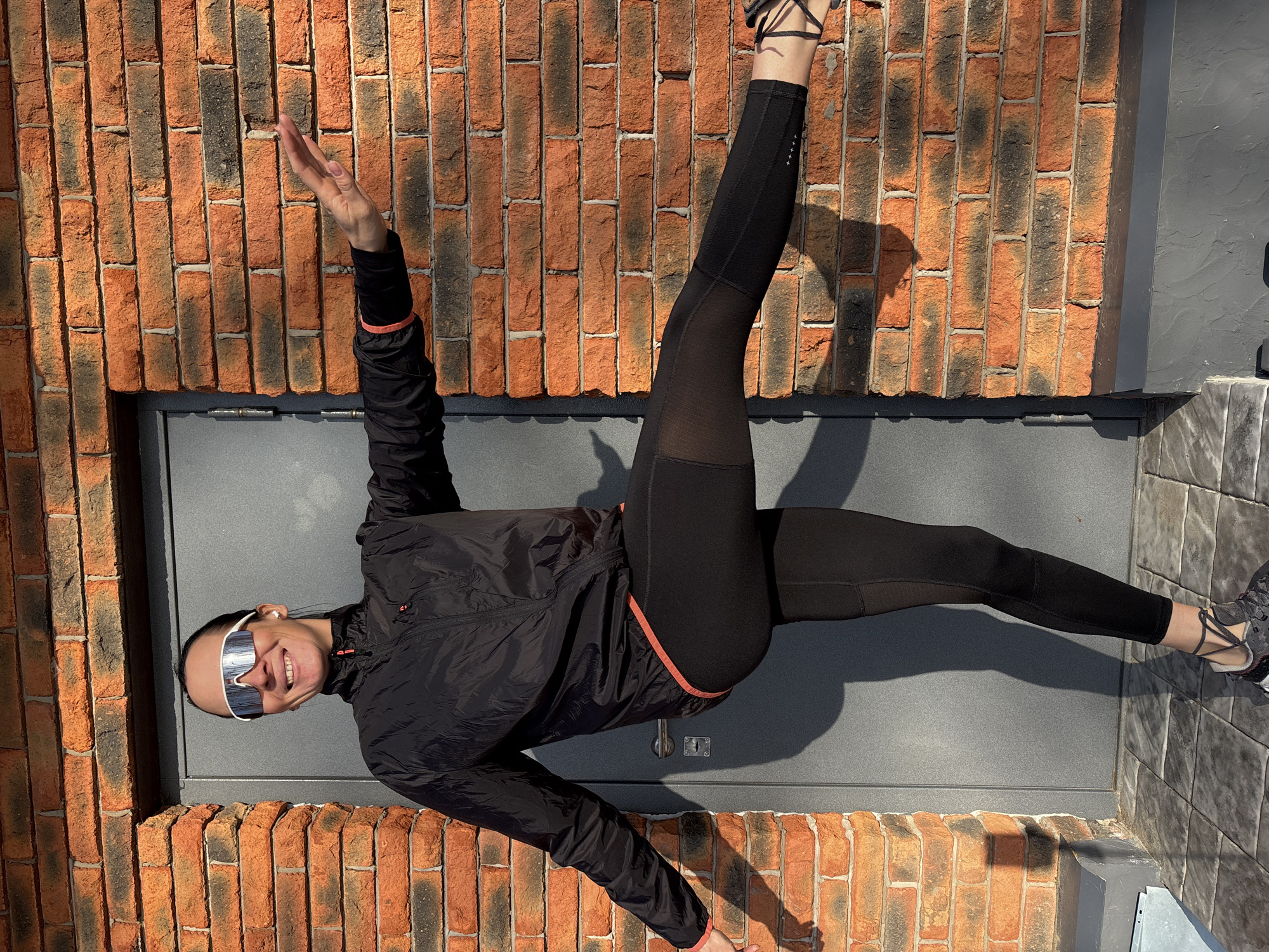 A woman wearing a black jacket with orange trim, black leggings with mesh panels, and white sunglasses is striking a pose outdoors. She is standing in front of a brick wall with a grey metal door or panel below it. She is wearing athletic shoes. The location is the city of Chaikovsky, Russia.