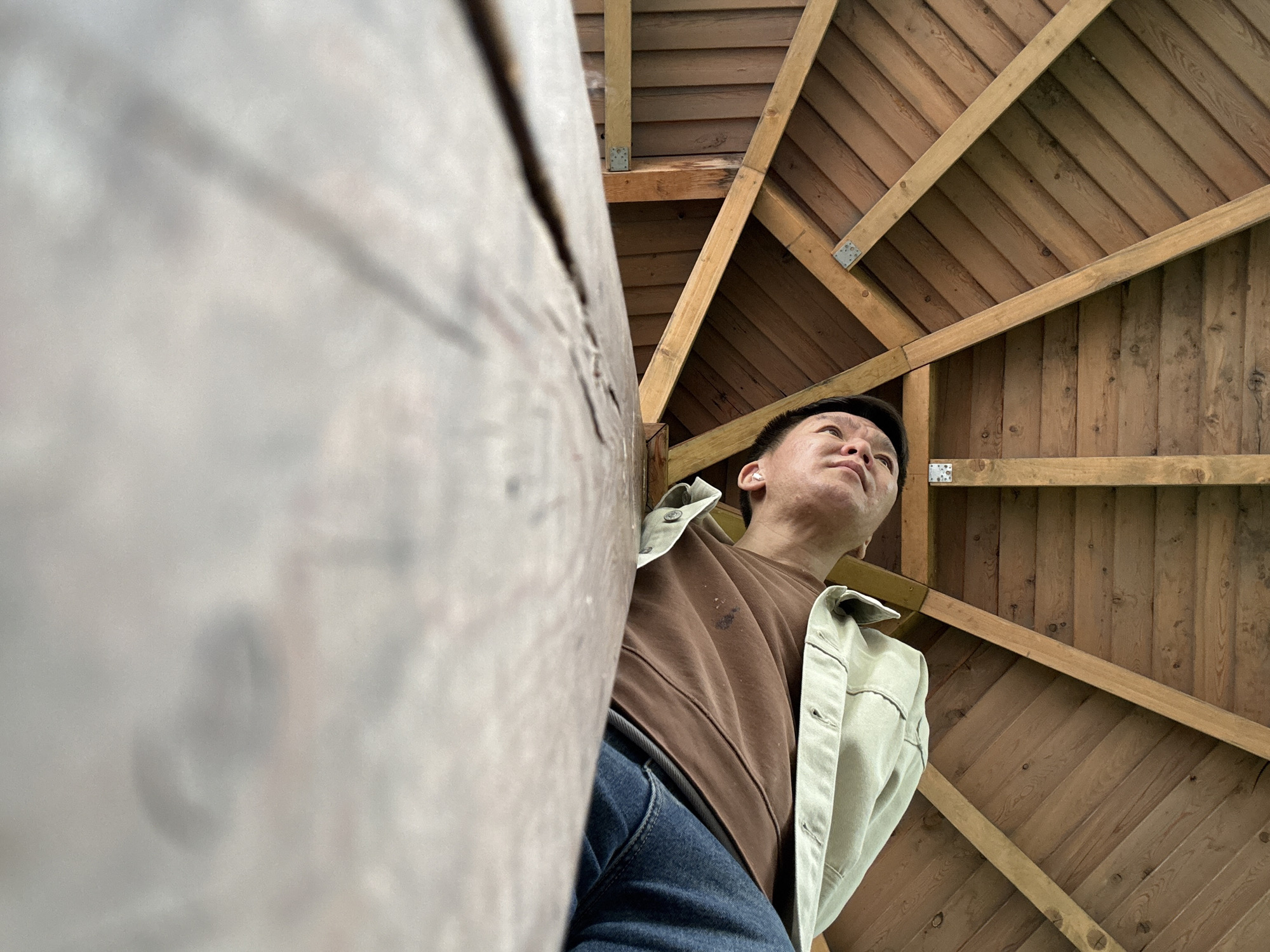 The image, taken from an extreme low-angle perspective, features a man looking upwards at a detailed wooden ceiling or roof structure. The man, who appears to be East Asian, occupies the lower right portion of the frame. He is dressed in a brown long-sleeved t-shirt, blue jeans, and a light-colored (possibly beige or light green) denim-style jacket. A white earbud is visible in his right ear. His short, dark hair and thoughtful expression suggest he is observing or contemplating the structure above him with curiosity.
The background reveals an elaborate wooden architectural design, likely the interior of a gazebo, shed, or timber-framed building in Moscow, Russia. Intersecting wooden beams and planks create a radial pattern, forming the roof or ceiling. Metal brackets are visible at the joints of some beams, securing the construction. The wood shows natural variations in grain and tone, indicating it is unpainted or stained. A large, out-of-focus wooden pillar or beam takes up a significant portion of the left foreground, adding to the sense of depth and emphasizing the low-angle shot. The lighting is soft and even, suggesting indirect daylight or an overcast day. No specific activity beyond observation is depicted, and no visible text is present in the image.