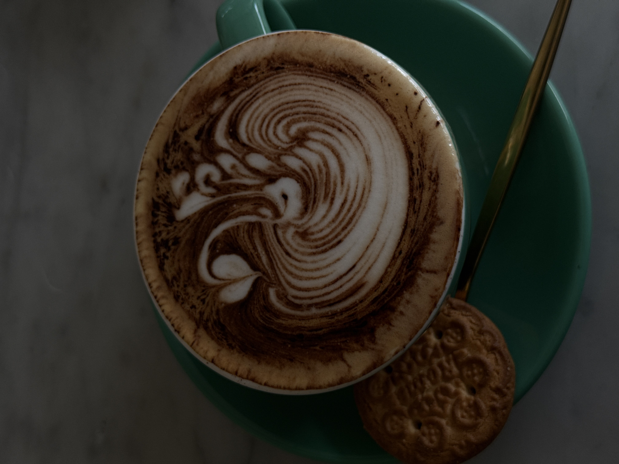 An overhead shot captures a beautifully crafted cup of latte art coffee, presented on a dark teal saucer. The coffee cup, also dark teal, is filled with a rich brown beverage topped with intricate white foam latte art, featuring a swirling, organic pattern that includes delicate, heart-like shapes. The rim of the coffee cup shows remnants of foamed milk.
Resting on the saucer beside the cup is a metallic, golden-colored spoon. Partially visible on the saucer, next to the coffee cup, is a round, light brown biscuit with an embossed, ornate pattern. The overall scene is set against a muted, dark grey background, possibly a stone or concrete surface, suggesting an indoor or shaded outdoor setting. The lighting is soft and subdued, contributing to a calm and contemplative mood. No discernible activity or people are present. There is no visible text in the image. This scene suggests a peaceful moment in North Kuta, Indonesia, perhaps at a cafe or a home.