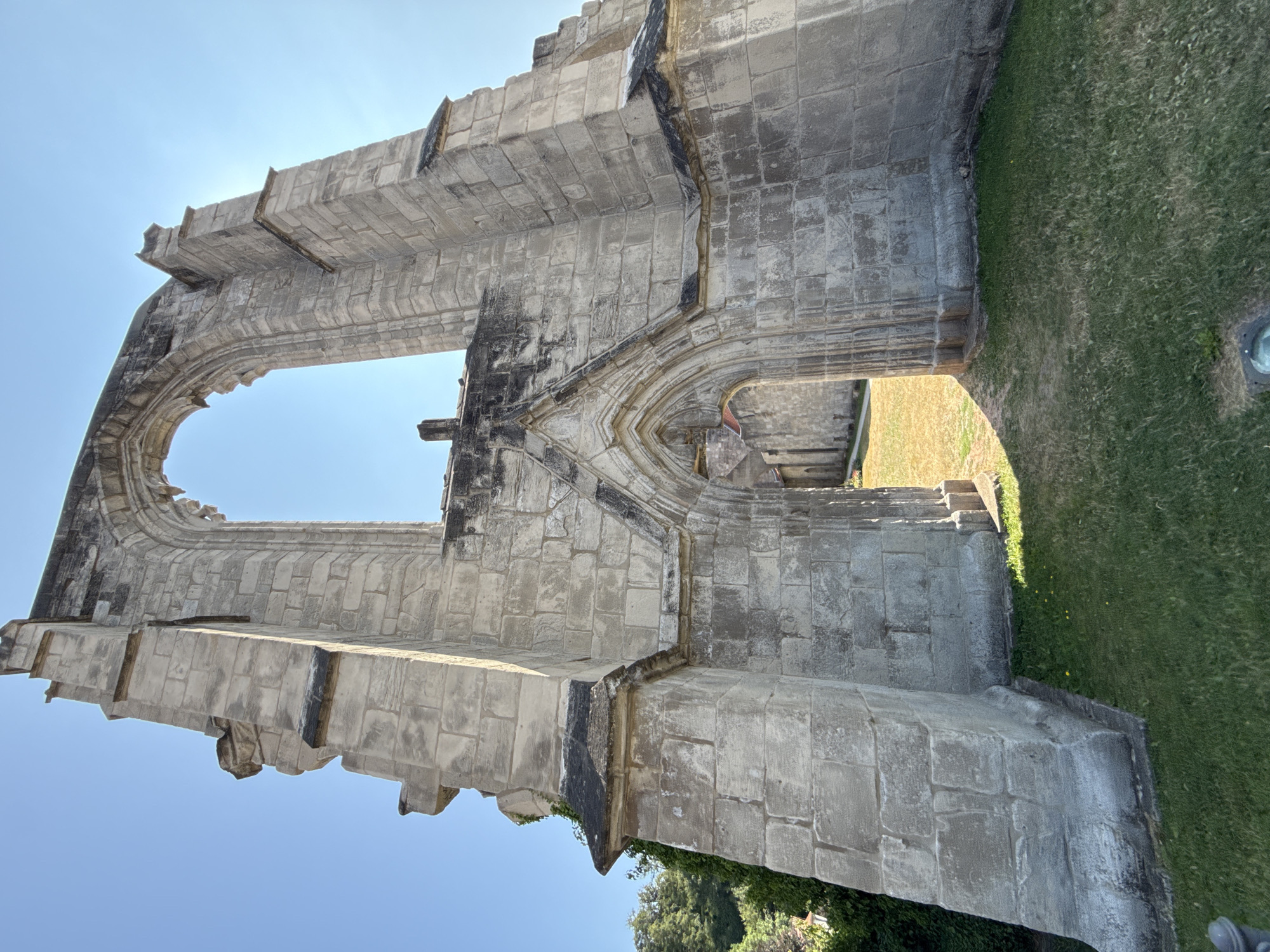The image depicts the ruins of the Walkenried Abbey in Walkenried, Germany. The focal point is a large, weathered stone archway, part of a Gothic structure that is largely in disrepair. The stonework is rough and shows signs of age and erosion. Sunlight streams through the open arch, illuminating the grassy area and a path leading to another section of the ruins in the background. The sky is clear blue. The foreground features a well-maintained lawn. To the left of the archway, ivy climbs the stone structure, and trees are visible further back. The scene is captured from a low-angle perspective, emphasizing the scale and grandeur of the ruins. There are no people or animals visible in the frame.