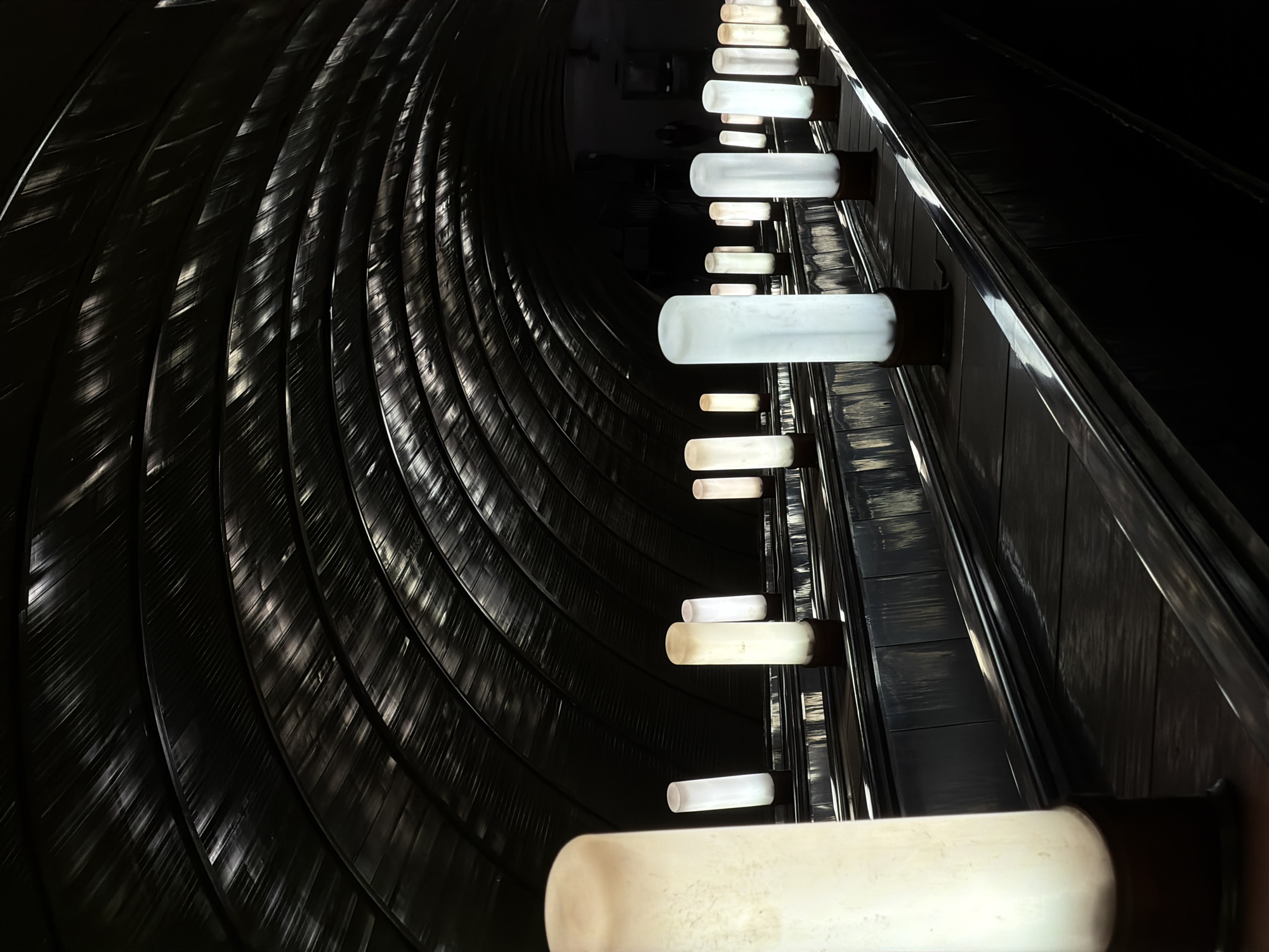 The image shows the interior of a dimly lit space, identified as the escalator tunnel at Park Pobedy station in the Moscow Metro. A curved wall with horizontal, closely spaced, reflective panels occupies the left side of the frame. A straight row of evenly spaced, cylindrical white lights is mounted on the right side of the frame, illuminating a dark, seemingly metallic, wall or structure. The perspective suggests looking upwards, likely from the base of an escalator. No people are visible.