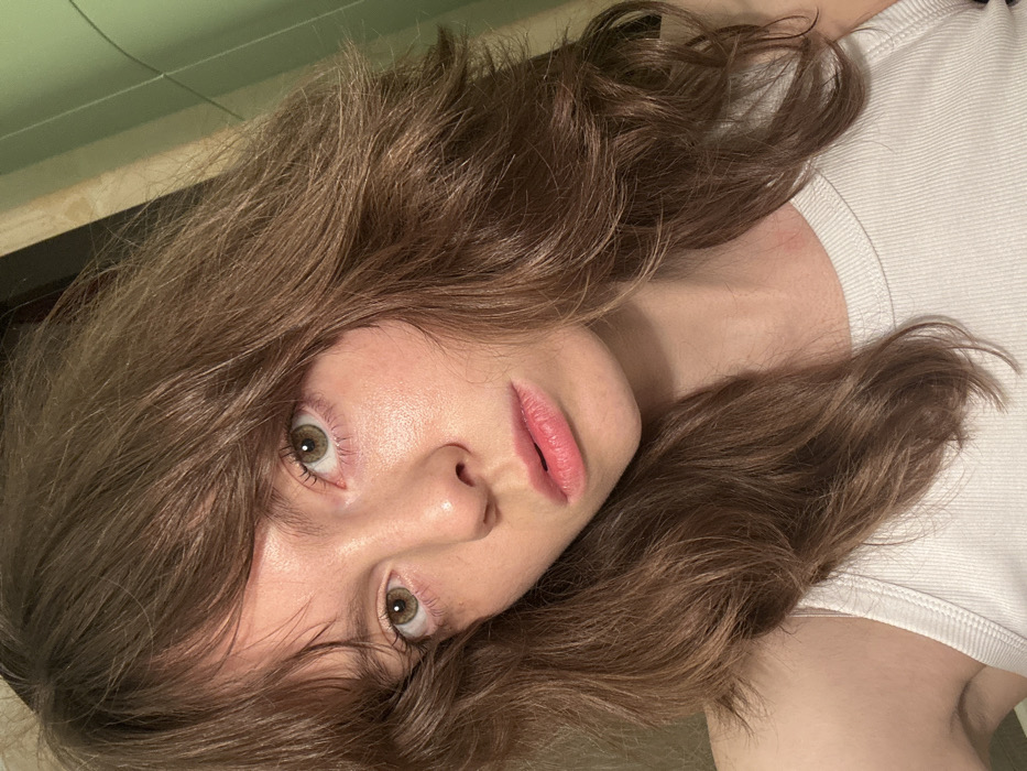 A close-up, head-and-shoulders portrait depicts a young adult female with fair skin, light brown, wavy, shoulder-length hair with bangs, and light green/hazel eyes. She wears a white ribbed tank top and maintains a neutral expression, looking directly into the camera. The background, largely out of focus, reveals an indoor setting with two distinct vertical panels: a textured, light beige or cream-colored surface on the left and a smooth, light green surface on the right. The specific location within the city of Moscow, Russia, is not identifiable from the visual evidence presented in the image.