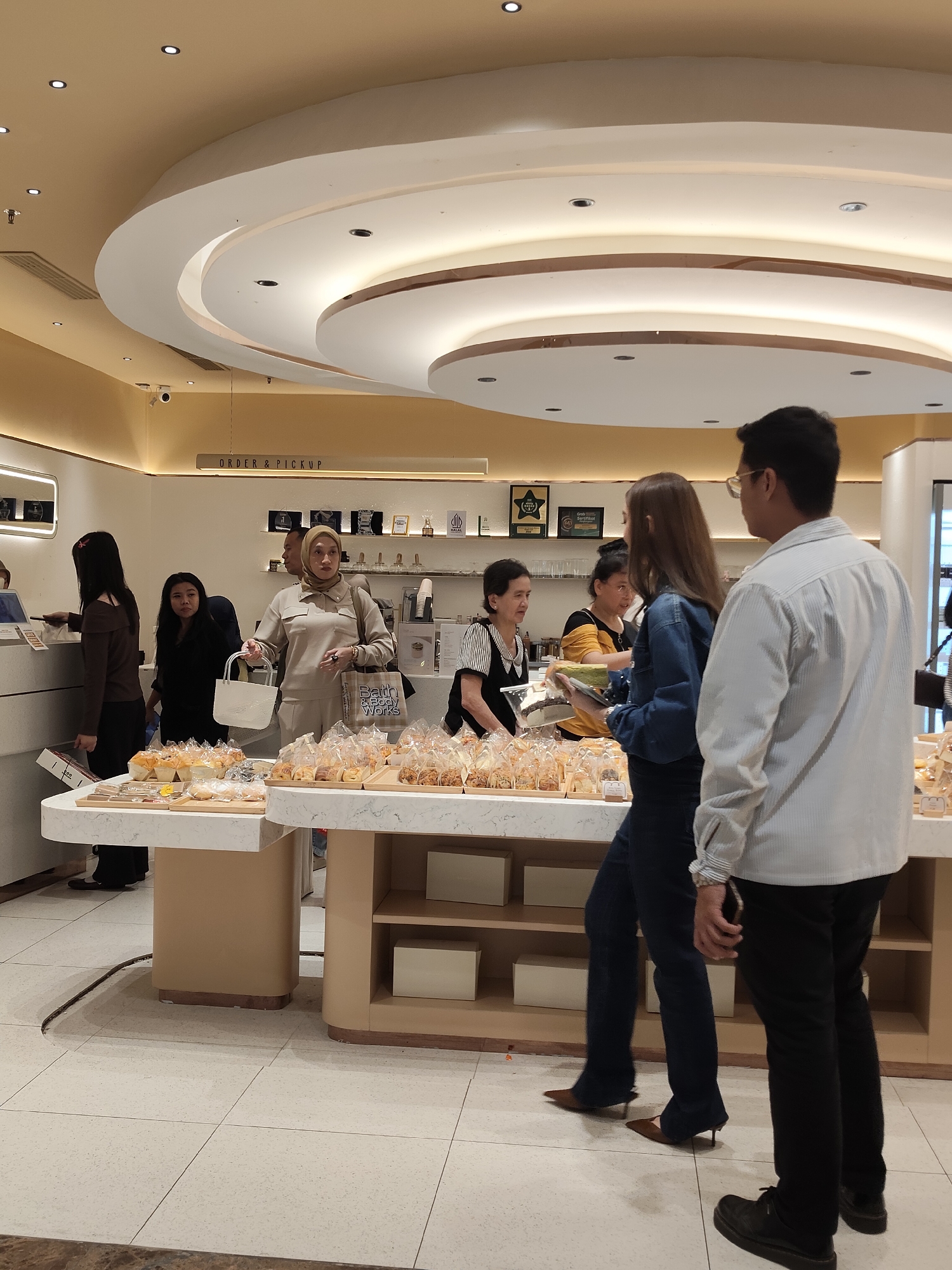 The image shows a bakery in Medan, Indonesia, with several customers and staff members present. The interior features a modern design with a curved ceiling and soft lighting. In the foreground, a display table is laden with freshly baked goods, many of which are individually packaged in clear plastic bags.
Several people are visible in the scene:
- A woman wearing a hijab and a beige outfit is holding a white shopping bag and a shopping bag from "Bath & Body Works." She appears to be a customer.
- A man in a light blue striped shirt and black pants is standing next to her, facing away from the camera.
- Another woman, with long brown hair, is wearing a blue denim jacket and dark pants, and is looking at a cake.
- Behind the counter, two women are interacting with customers. One appears to be a staff member with dark hair, and the other has lighter hair and is wearing a black and yellow striped shirt.
- To the left of the frame, another woman with dark hair is standing near a counter, possibly a cashier.
The environment suggests a retail setting. Shelves behind the counter display various items, and a sign reading "ORDER & PICKUP" is visible on the wall. The floor is tiled. The overall impression is of a busy but well-organized bakery.