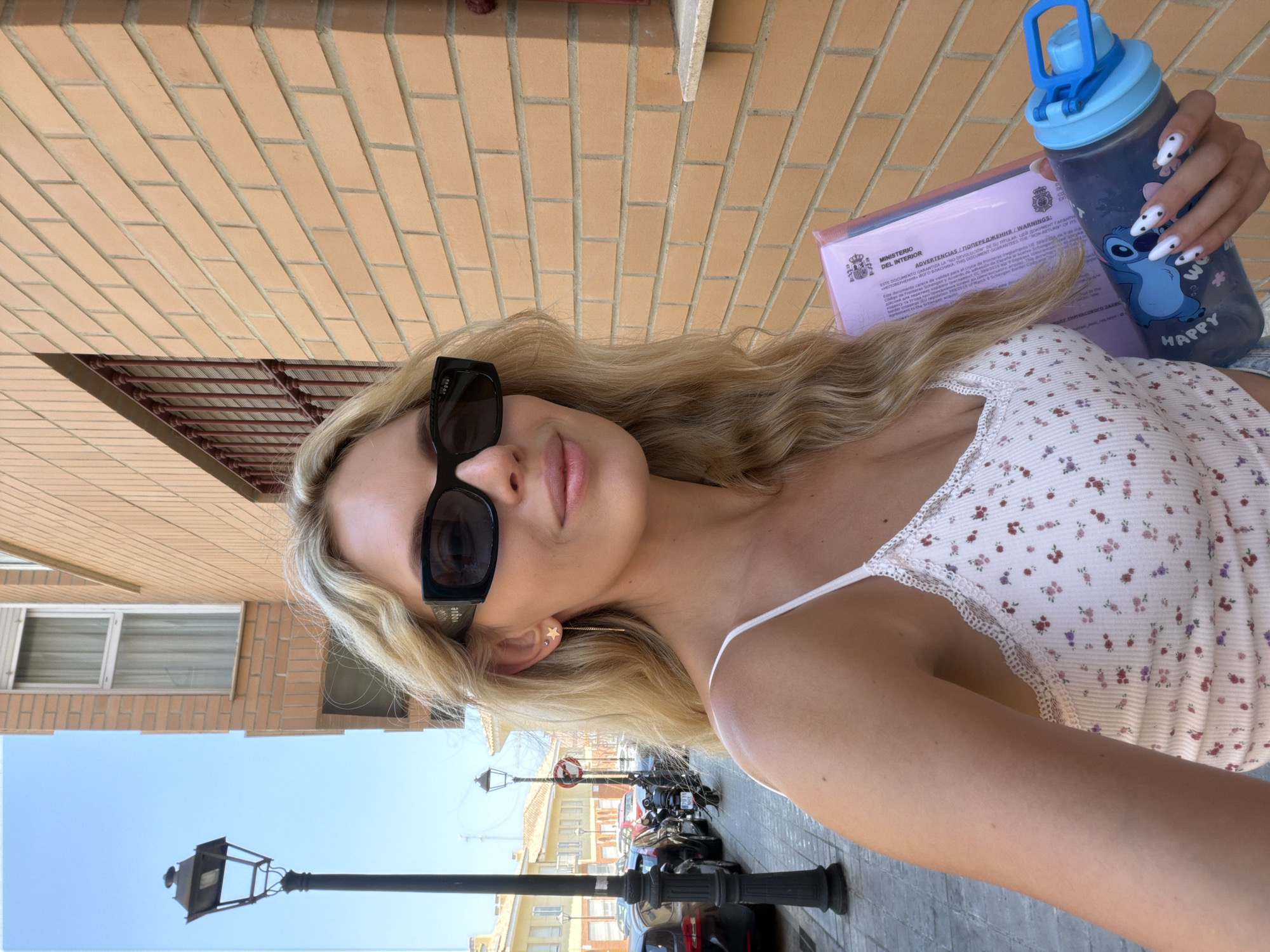 The image shows a young blonde woman with wavy hair, wearing black oversized sunglasses and a white floral print tank top. She is holding a pink folder and a blue water bottle with a Stitch cartoon character design. The woman is standing on a sidewalk next to a brick building. In the background, there is a street with parked cars and a lamppost. The scene appears to be in Benimàmet-beniferri, Spain, indicated by the style of architecture and the presence of Spanish text on the folder. The lighting suggests it is daytime, likely sunny.