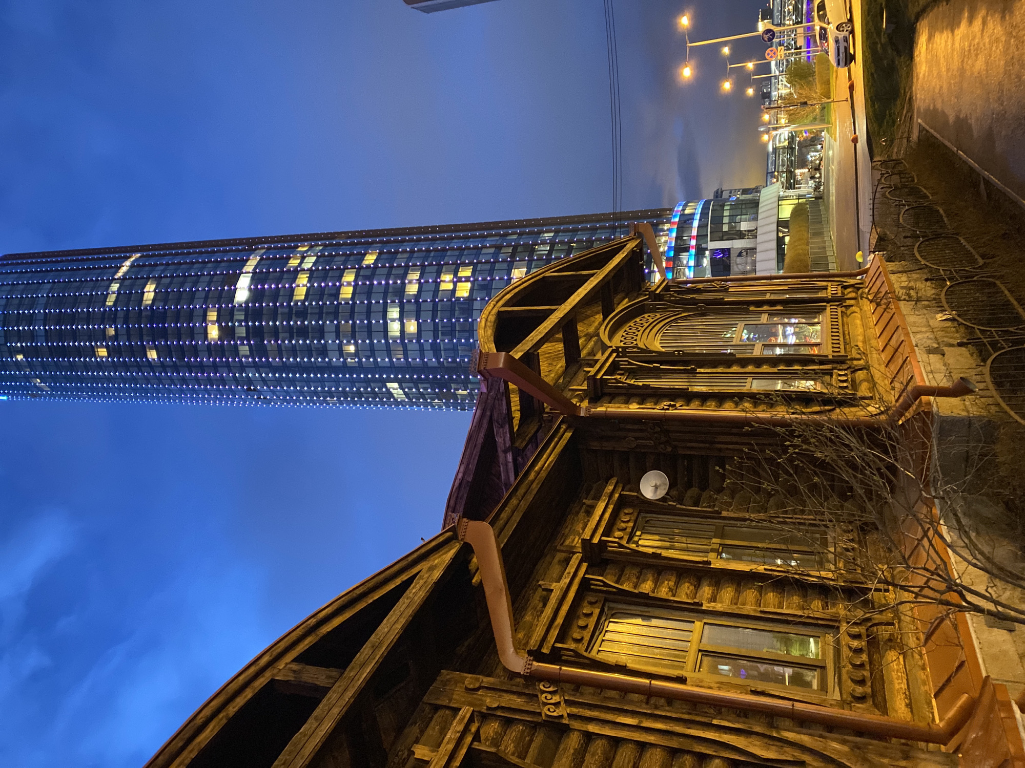 This evening photograph captures a striking juxtaposition of old and new architecture in an urban setting, identified as Yekaterinburg, Russia. The image is taken at a low angle and rotated, giving a dynamic perspective of the twilight scene.
In the foreground and right side, an ornate, rustic wooden building stands prominently. Its dark timber facade features multiple windows with intricate frames, carved decorative elements, and visible downspouts and gutters. A small, white satellite dish is affixed to one side. The building's windows emit a warm, golden light, suggesting interior activity. A bare tree branch with fine twigs partially obscures the right side of this structure.
Dominating the midground and left side of the frame is a tall, modern skyscraper. Its curved glass facade is illuminated by countless small, purple-blue lights arranged in a grid pattern, alongside various yellow-lit windows. This creates a contemporary, almost futuristic appearance, contrasting sharply with the traditional wooden building.
Beyond the wooden building and to the right, a street scene unfolds with multiple streetlights casting a warm glow onto a paved path or sidewalk. Further back, another modern, glass-fronted building, possibly a commercial center, is visible with reflections of red and blue lights. A white car is partially visible on the street, and indistinct traffic signs are present on poles.
The sky is a deep, dark blue, indicating late dusk or early night, with hints of cloud cover. The overall impression is one of a city where historical structures coexist with modern development, bathed in the artificial lights of evening. No specific activity or event is occurring, but the illuminated buildings suggest a lively urban environment. No legible text is visible.