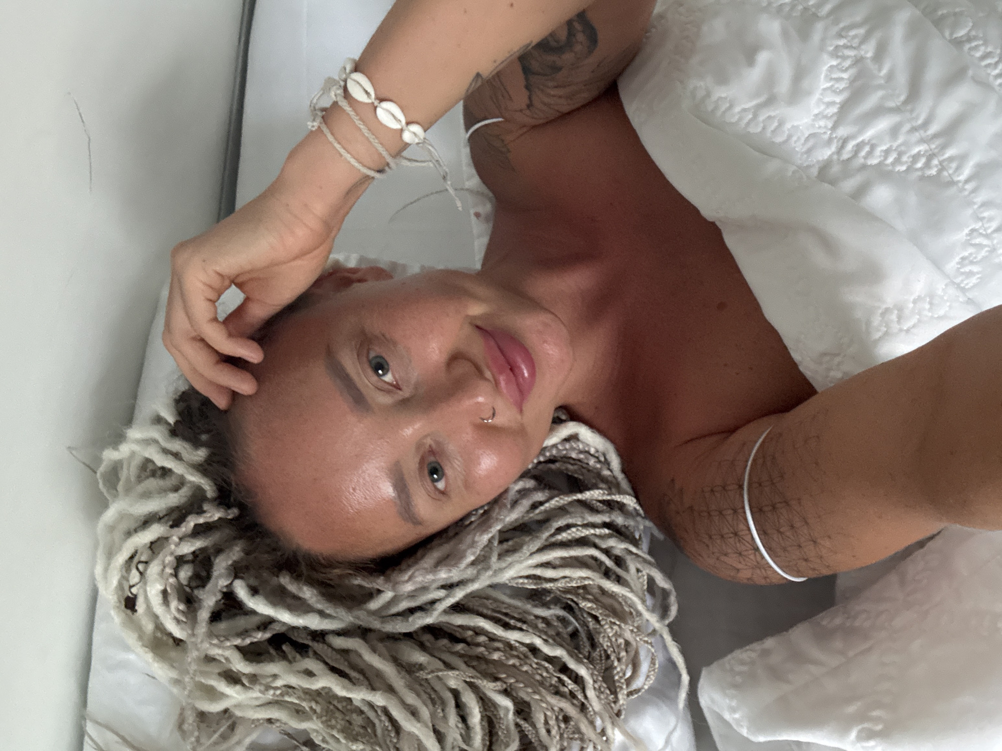 A woman with blonde dreadlocks and a nose ring lies on a white bed, looking at the camera with a slight smile. She is wearing a white top and bracelets on both arms. Her left arm has a tattoo. The background is a white wall and bedding. The location is not specified.