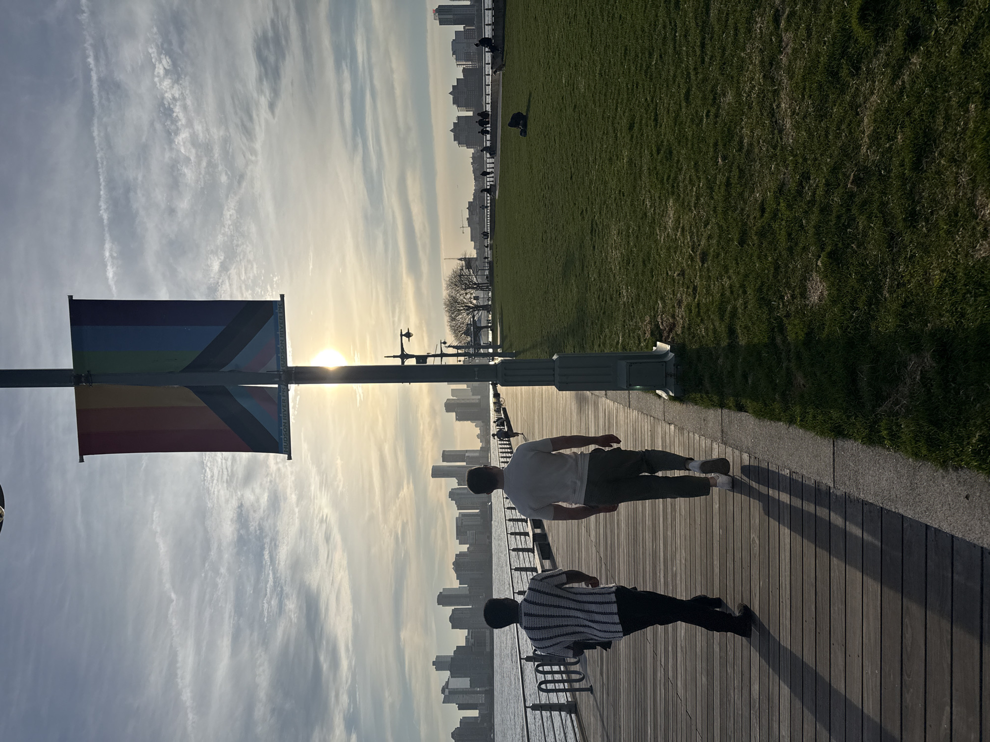 The image depicts a scene along the Hudson River in New York City. Two people walk on a wooden boardwalk, one in a white shirt and one in a black and white striped shirt. A rainbow flag hangs from a pole. Beyond the boardwalk is a stretch of grass with several people sitting on a railing. In the background is the skyline of buildings, partly obscured by a sun flare. The sky is lightly cloudy.