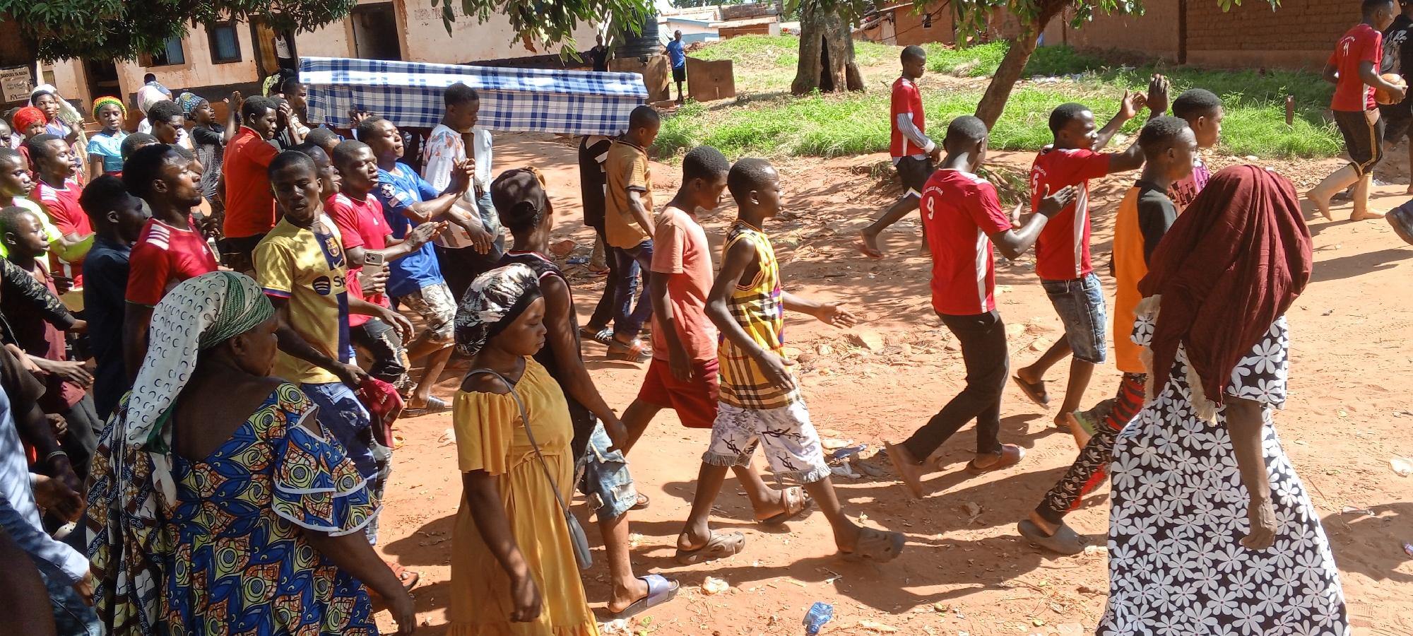 A large group of people, mostly African, is seen walking on a red dirt ground in Ngalula, Democratic Republic of the Congo. The crowd consists of men, women, and children dressed in casual clothing, including T-shirts, dresses, shorts, and patterned fabrics. Some are carrying a mattress with a blue and white checkered pattern, suggesting they are moving or transporting it. Many members of the group are looking in the same direction, with some raising their hands, possibly chanting or cheering. Buildings and trees are visible in the background. The image suggests an outdoor scene, with strong sunlight casting shadows on the ground.
