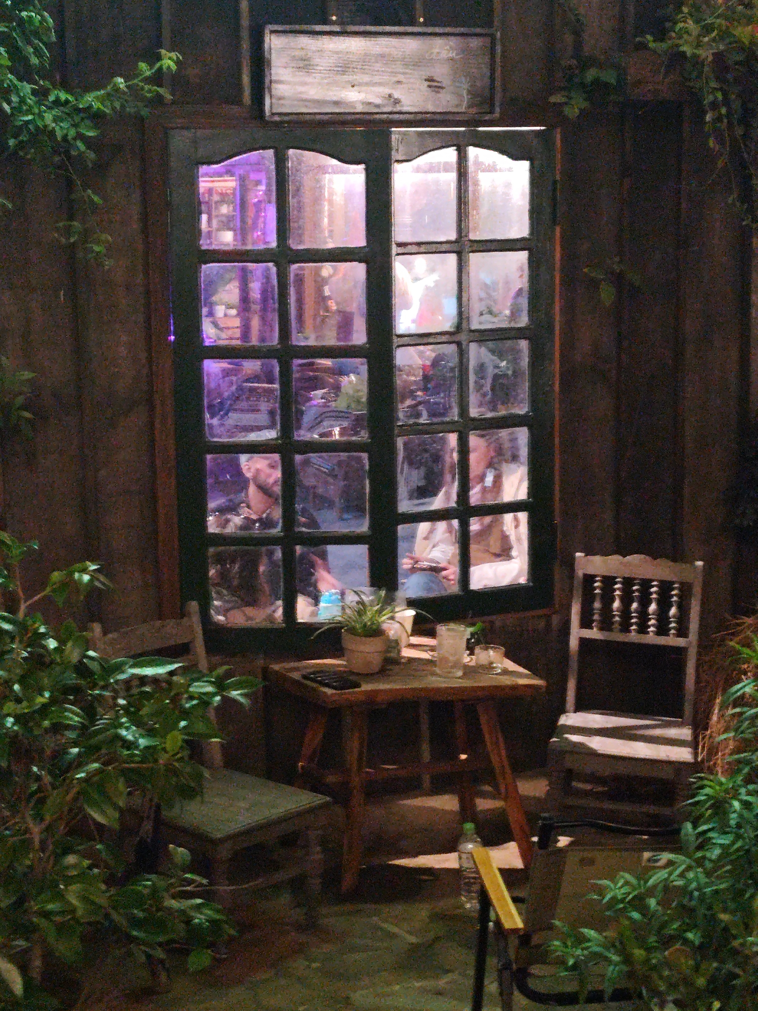 A view from a café in Xuan Truong - Da Lat, Vietnam, focuses on a small wooden table flanked by two wooden chairs, situated near a multi-paned window. The window reflects the interior of the café, where two individuals are visible: a man wearing a patterned shirt, and a woman in a light-colored top and vest. On the table, there is a small potted plant, a drinking glass, and an unidentified object. A water bottle lies on the floor near the table. The scene is framed by green foliage, suggesting a natural or garden-like setting. Above the window, there's a wooden sign. The lighting within the café appears to be purple-hued.
