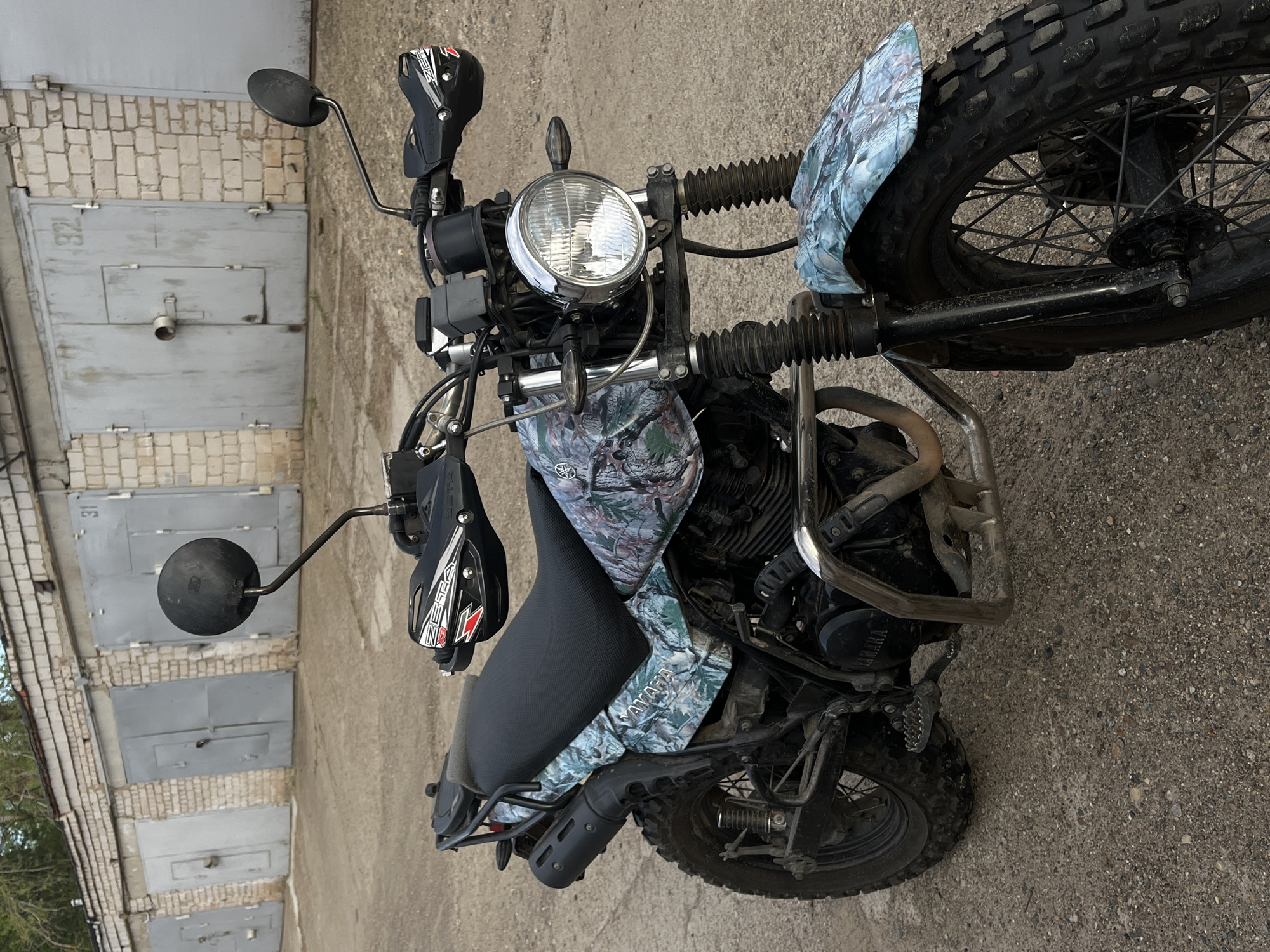 The image depicts a Yamaha motorcycle parked in front of a row of garage doors in the city of Chaikovsky, Russia. The motorcycle is customized with a camouflage pattern on the front fender, fuel tank, and side panels. It features a round headlight, black seat, aftermarket handguards, and knobby tires. The garage doors are painted gray and show signs of wear and tear. The ground is asphalt.
