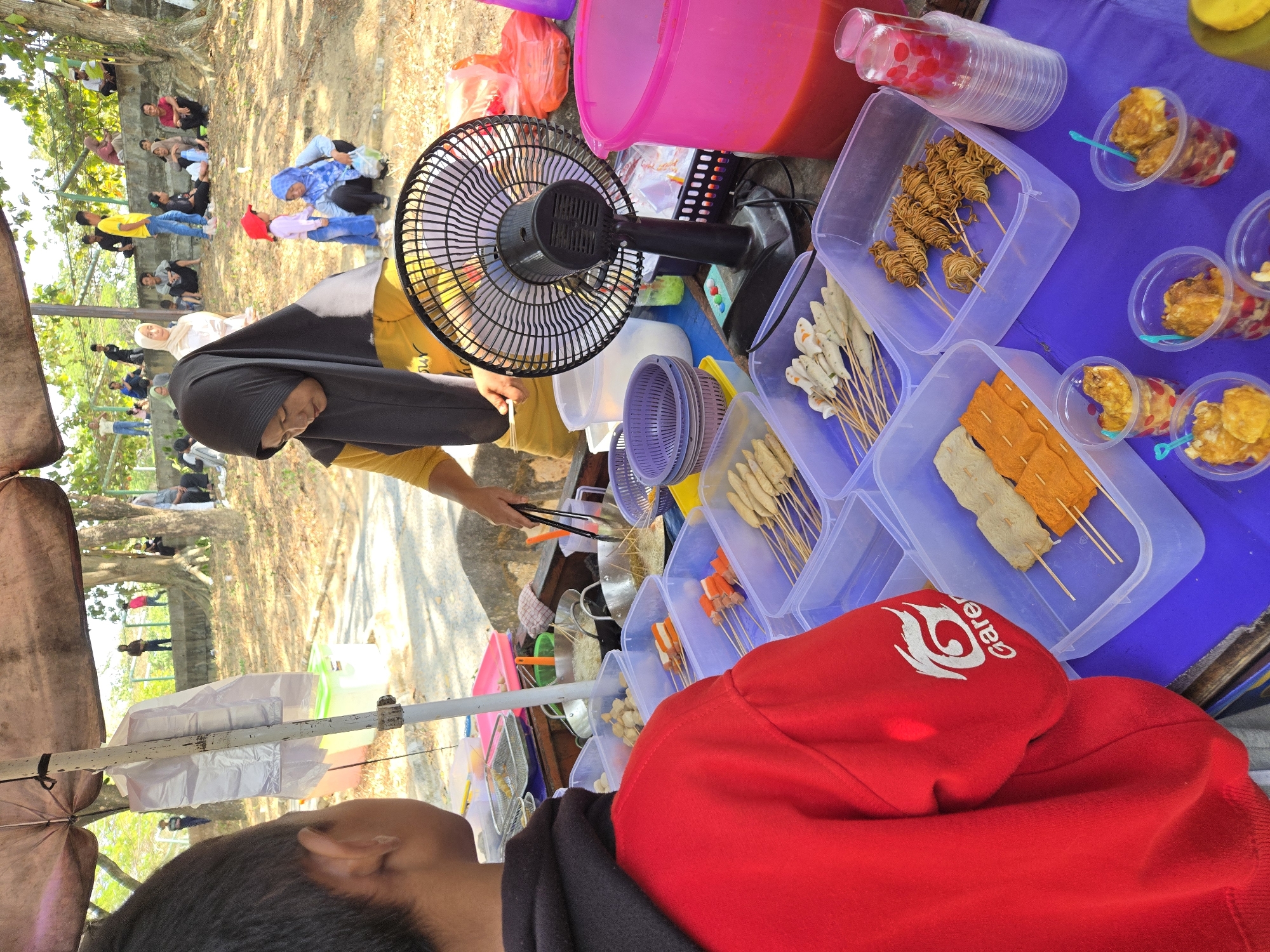 In Sungailiat, Indonesia, a street food vendor with dark hair and wearing a black hijab and a yellow shirt is serving customers. She is using tongs to cook food in a pan, and a young person in a red hooded jacket with a white "Garena" logo on the sleeve is standing in front of the stall, looking towards the vendor. The stall has several plastic containers filled with various skewers of food, including what appear to be fish cakes, sausages, and fried noodles. There's also a black electric fan on the table. In the background, people are gathered on a sandy area with a concrete wall and trees. The scene appears to be an outdoor market or gathering.