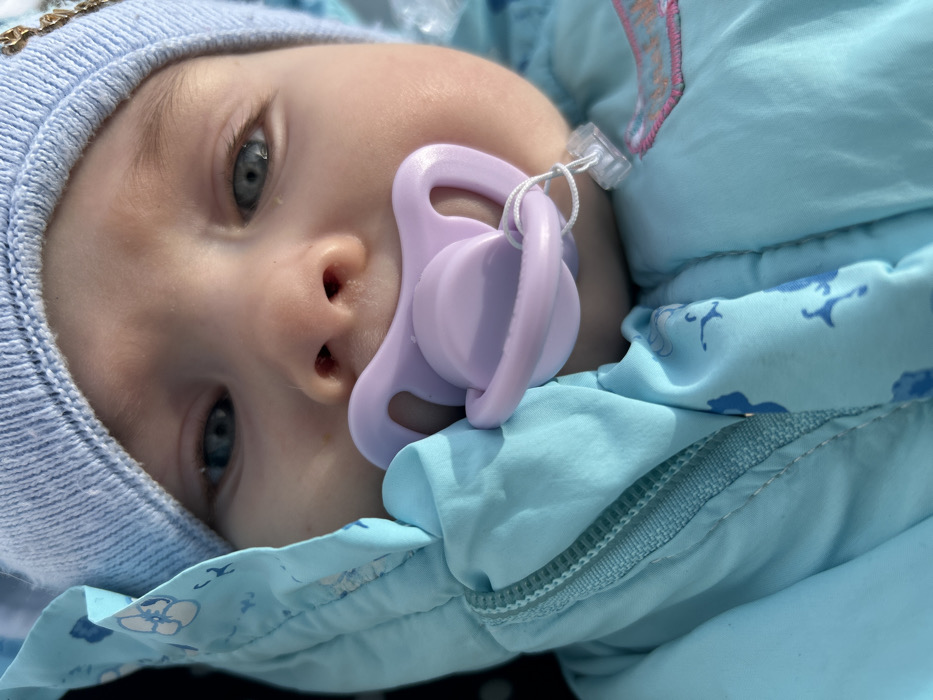 A close-up, high-angle shot captures an infant outdoors in Chaikovsky, Russia. The infant, displaying fair skin and blue eyes, is wearing a light blue knitted hat and a light blue hooded winter garment, possibly a snowsuit, featuring faint darker blue animal patterns. A silver-colored zipper is visible on the left side of the garment. A light purple pacifier is inserted into the infant's mouth, connected by a thin white string to a partially visible clear plastic clip near the lower right. Strong natural light illuminates the scene, casting soft shadows. A small section of a gold-colored chain or ornament is faintly visible near the upper right edge, adjacent to the hat. The infant's attention is directed towards the camera.