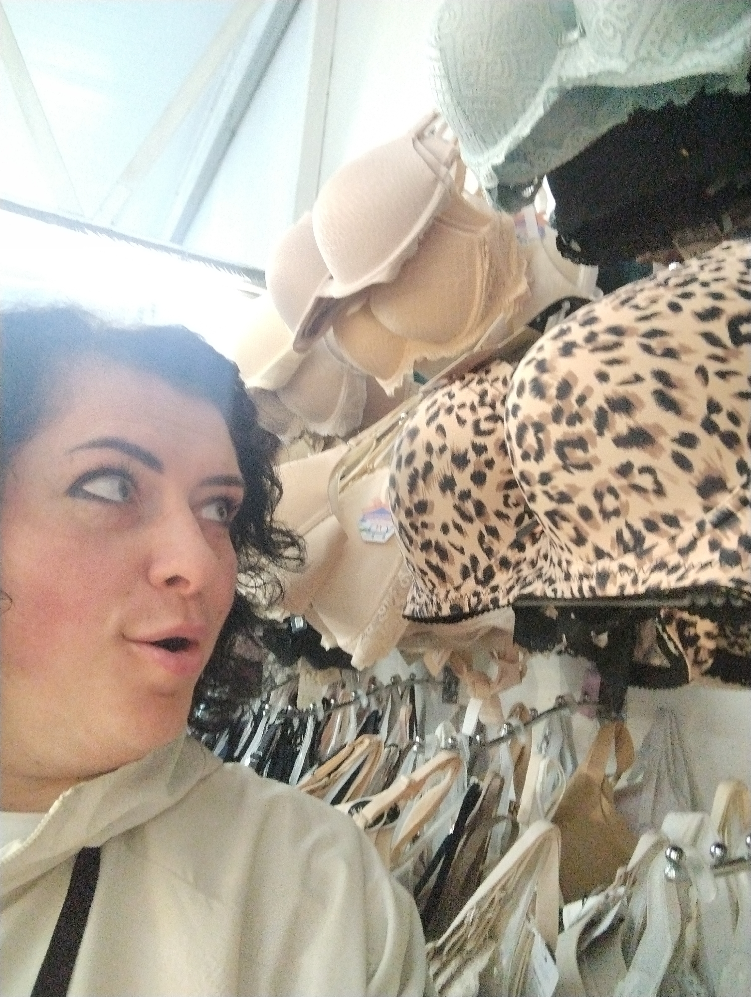 A woman with dark curly hair and a tan jacket is standing in front of a rack of bras. The bras include tan, black, and leopard print designs. The environment appears to be a clothing store. There is no evidence to suggest that this image was taken in the city of Votkinsk, Russia.