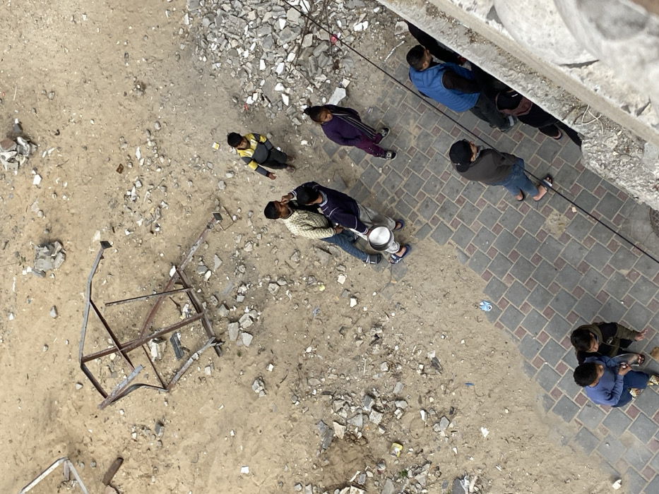 An overhead image depicts a street-level scene in Khan Yunis, Palestinian Territory. The ground is divided into two distinct areas: the majority of the frame, in the upper and central portions, is an expanse of light brown, dry dirt heavily interspersed with concrete rubble, fragmented blocks, and scattered debris. In this dirt area, a large, rusted, and bent metal framework, possibly a component of a structure or large object, rests on its side in the upper-left quadrant. A concentrated pile of concrete fragments is located on the right side of the dirt expanse, adjacent to the paved area.
The lower portion of the image shows a sidewalk constructed from interlocking grey and reddish-brown pavers arranged in a grid pattern.
Nine individuals are visible. On the dirt area, four individuals are present. Two young males stand centrally: one wears a light, patterned long-sleeve shirt and blue jeans; the other, in a dark blue long-sleeve top and grey pants, holds a large, empty-appearing metal pot by his left side, with his right hand extended towards the first male's face. A younger child in a yellow and grey striped long-sleeve top stands behind them, looking right. A female child in a dark purple long-sleeve top and dark pants stands to the right, near the rubble pile.
On the paved sidewalk, five individuals are seen. An adult male in a dark jacket, blue jeans, and flip-flops stands looking towards the dirt area. To his left, a young child in a light blue long-sleeve top and dark pants stands next to an adult or older youth wearing a dark jacket. On the far right of the sidewalk, partially obscured by a concrete barrier, are two more individuals: an adult male in a blue vest over a dark shirt, and another person largely obscured behind him. A dark cable or wire runs vertically along the concrete barrier on the far right.