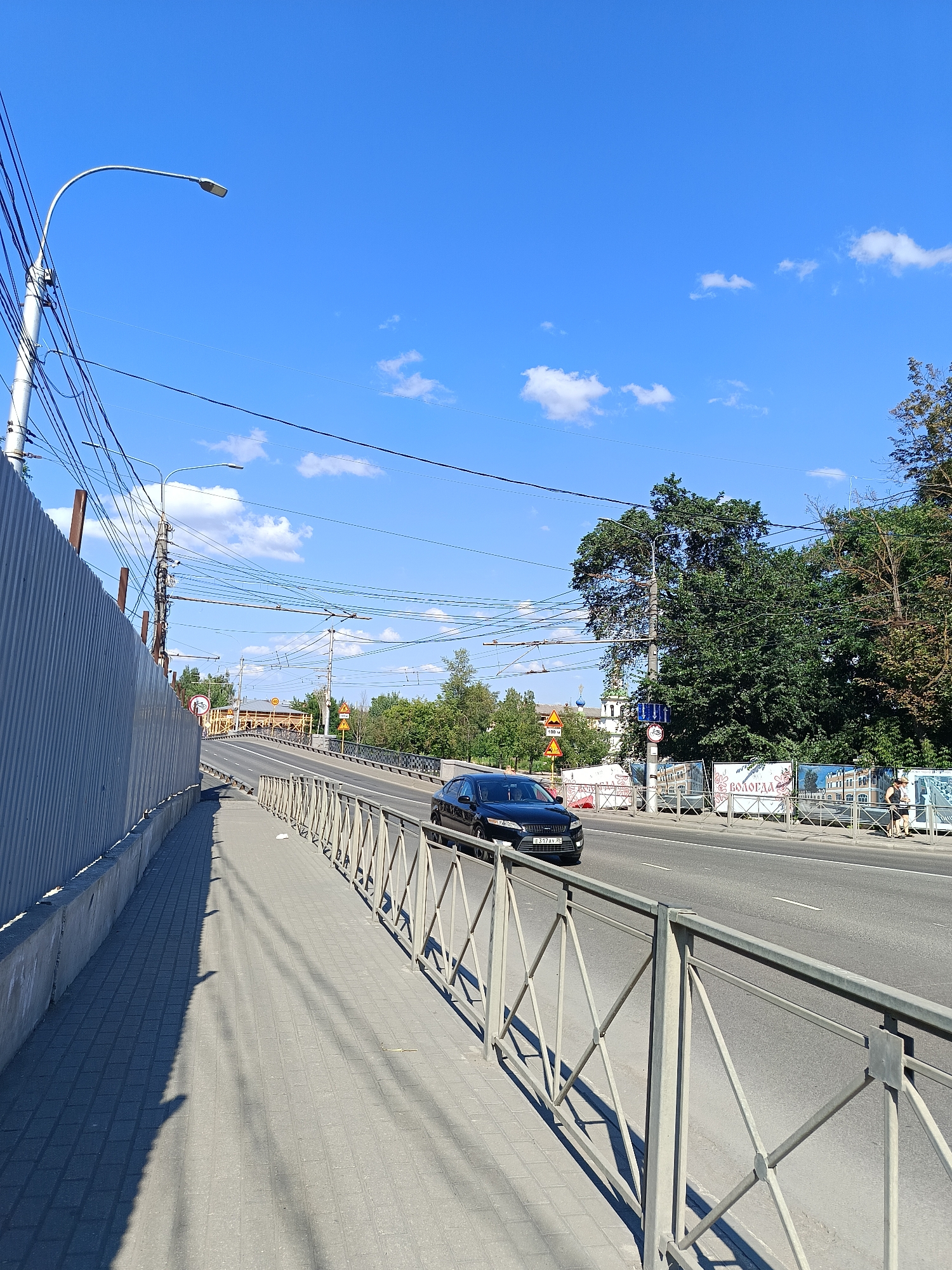 The image captures a bright, sunny day in an urban environment, likely a city, given the presence of a road, sidewalk, power lines, and buildings. The perspective is from a sidewalk looking towards a road where a dark-colored car is driving. The sidewalk is paved with gray tiles and is bordered by a metal railing. A tall, ribbed gray metal fence runs along the left side of the sidewalk. Overhead, a complex network of power lines and streetlights crisscrosses the sky, which is a vibrant blue with scattered white clouds. Trees are visible in the background, and some of them are quite large and leafy. There are also some banners and signs visible on the right side of the road, one of which appears to display text in Russian and images of buildings. Two people are walking on the sidewalk on the right side of the road. The overall impression is of a typical city street scene on a clear day.