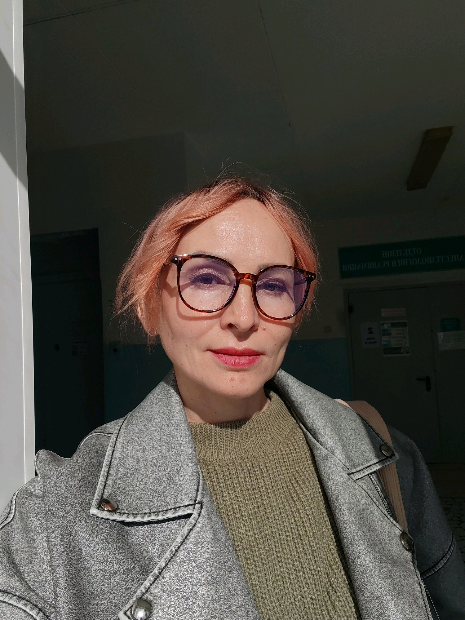 A close-up, head-and-shoulders selfie of a woman with short, pink-dyed hair and glasses. She is wearing a grey leather jacket over a green knitted sweater. The background appears to be an indoor hallway, possibly in a medical facility, with a green sign above a door. A hint of sunlight is illuminating the woman's face and hair, casting shadows. The overall lighting suggests it is daytime.