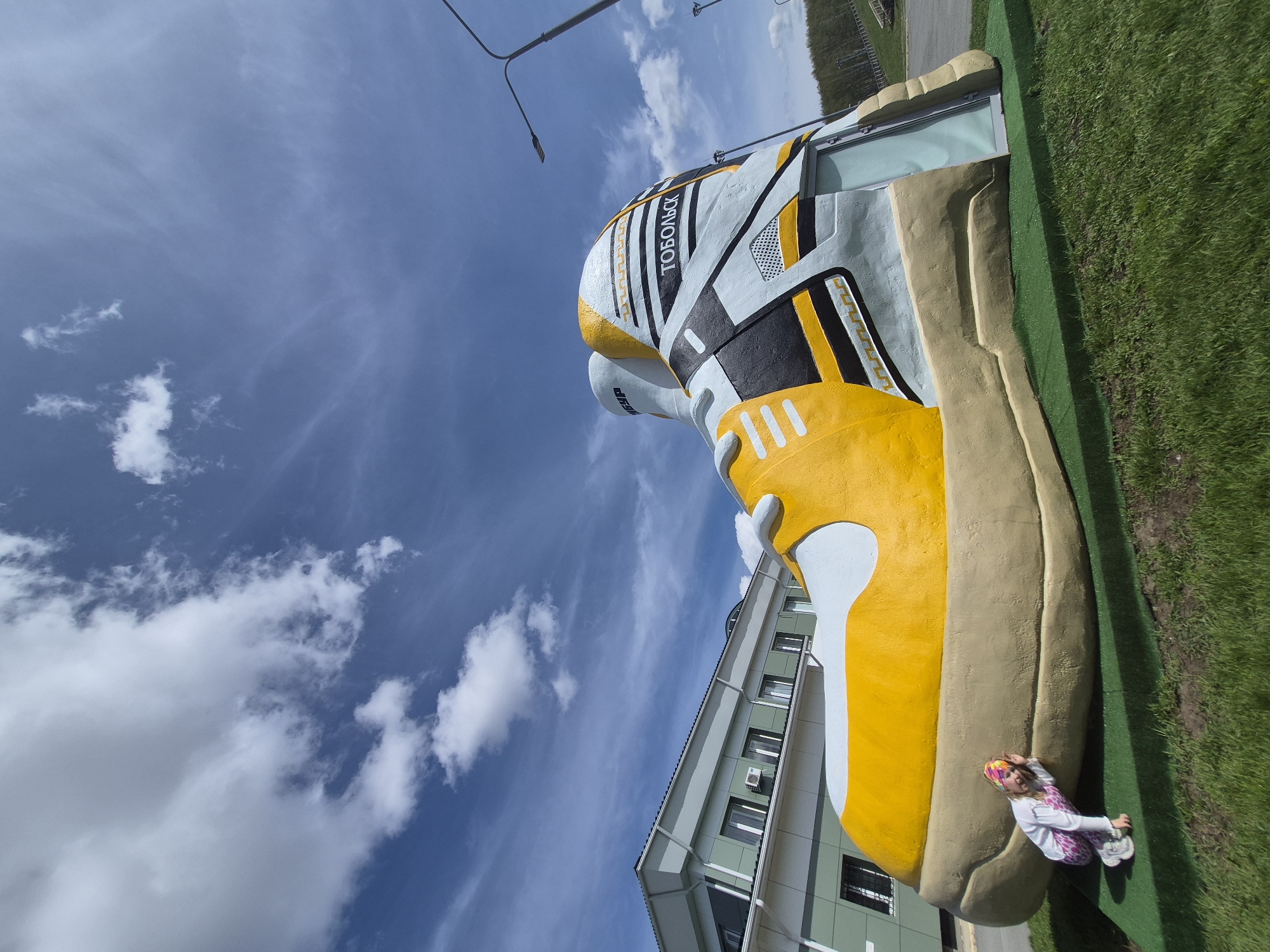 The image shows a large, colorful sculpture of a sneaker, situated in what appears to be an urban area, likely in Tobolsk, Russia, as the word "ТОБОЛЬСК" is written on the sneaker. The sneaker sculpture is primarily yellow, white, and black with a tan sole. It stands adjacent to a green lawn, a building, and a lamppost. A young child wearing pink pants, a white top, and a headband is sitting at the base of the sneaker, seemingly interacting with the sculpture. The sky is a bright blue, filled with puffy white clouds.