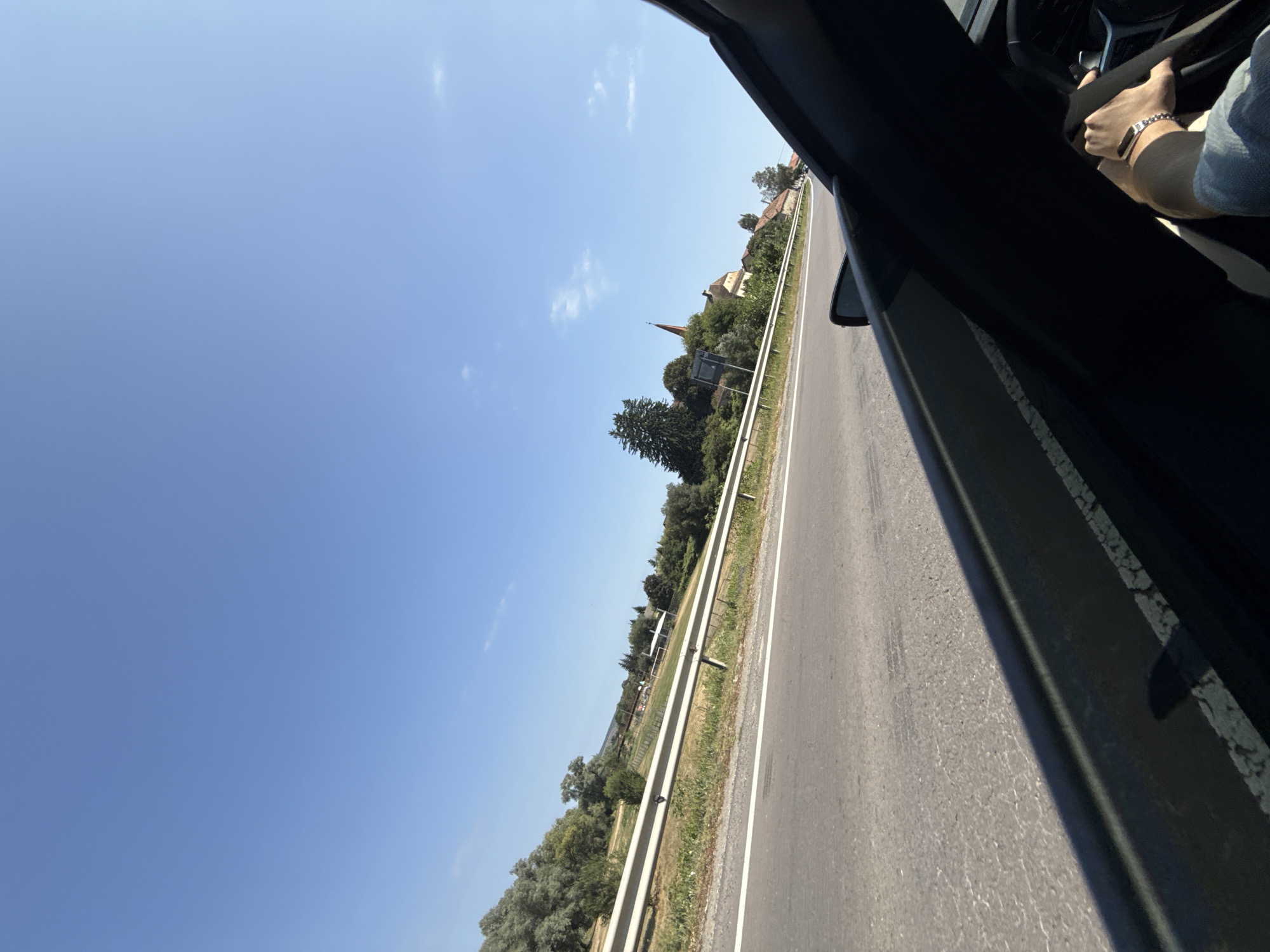 The image captures a view from the passenger seat of a car traveling along a road, likely near Șercaia, Romania, given the presence of a small village or town with a church spire in the distance. The road is paved and marked with white lines, indicating a multi-lane highway or a major road. A metal guardrail runs along the side of the road, separating it from a grassy embankment with trees and vegetation. In the background, a cluster of buildings, including a prominent church with an orange spire, are visible amidst a verdant landscape. The sky is a clear, bright blue with a few wisps of clouds. The interior of the car is partially visible, showing a hand on the steering wheel, suggesting the driver is operating the vehicle. The overall scene depicts a sunny day during travel through a rural or semi-rural area.