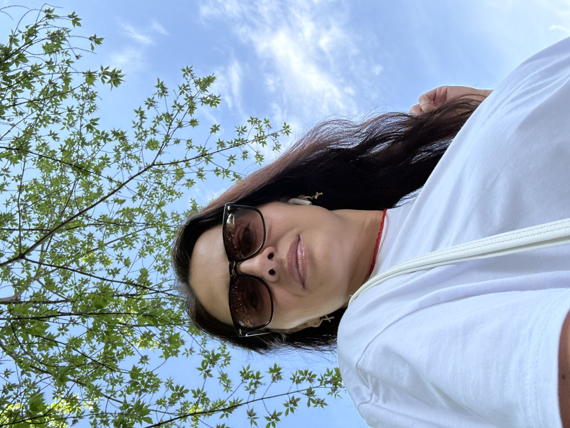 A woman wearing sunglasses and a white t-shirt stands beneath a tree with green leaves against a backdrop of a partly cloudy blue sky. The woman's face is centered in the frame. She has long, dark hair and is wearing wireless earphones. The photo is taken from a low angle, looking up. The location is likely in the city of Chaikovsky, Russia.