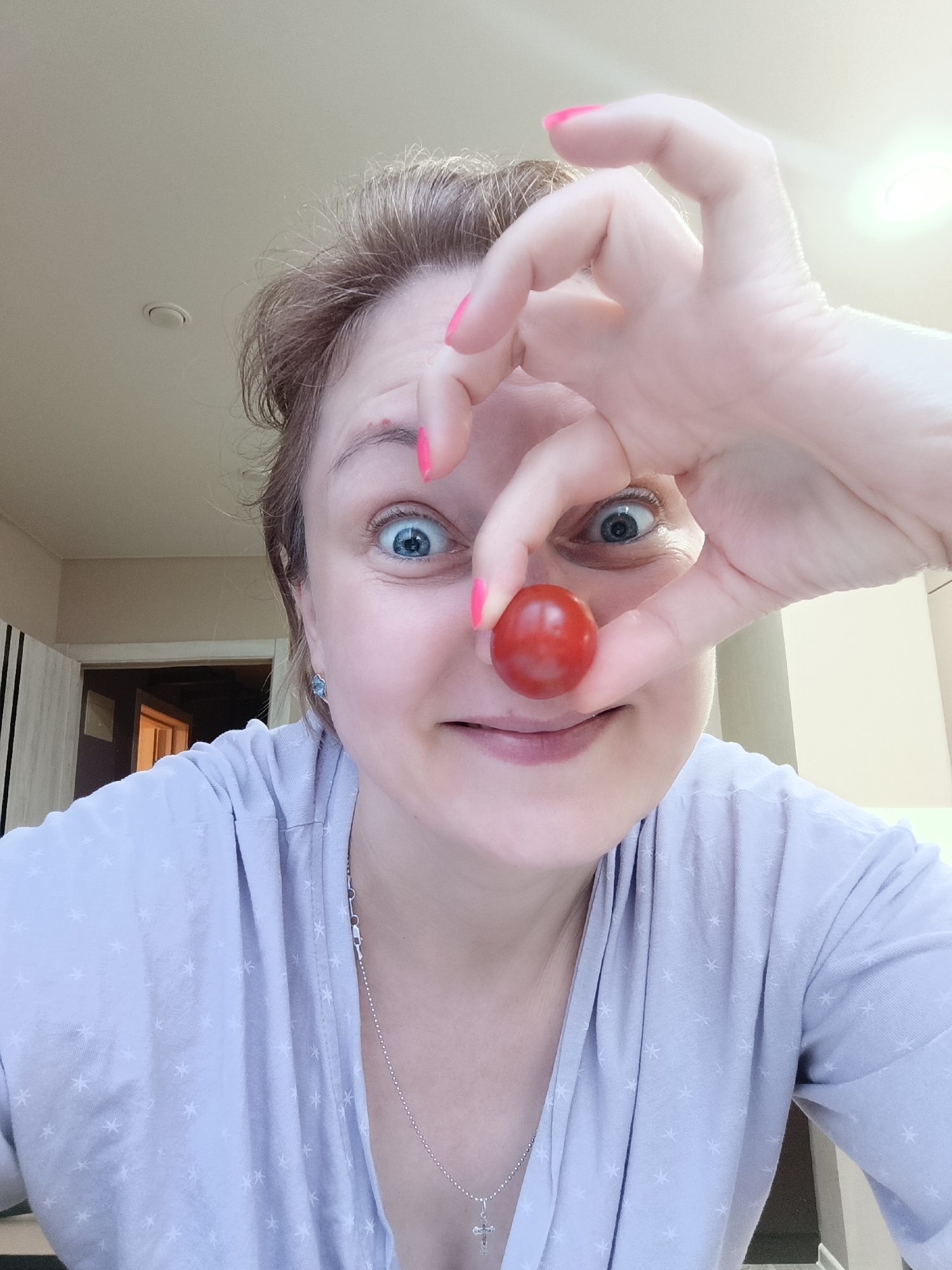 A woman is captured in a close-up selfie, playfully holding a small, red cherry tomato between her thumb and forefinger directly in front of her nose, creating the impression of a clown nose. She looks directly at the camera with wide, light blue eyes and a subtle smile, conveying a sense of humor. Her light brown hair frames her face.
She is wearing a light-colored garment, possibly light blue or lavender, adorned with a repeating pattern of small white stars. A silver chain necklace with a small cross pendant is visible around her neck. Her fingernails are painted with bright pink polish.
The background suggests an indoor setting, likely a home. A doorway is visible on the left leading into another room. The upper part of the image shows a light-colored ceiling with a recessed light fixture. The lighting is bright, indicating it is likely daytime indoors. No visible text is present in the image. The overall impression is one of a lighthearted and spontaneous moment.