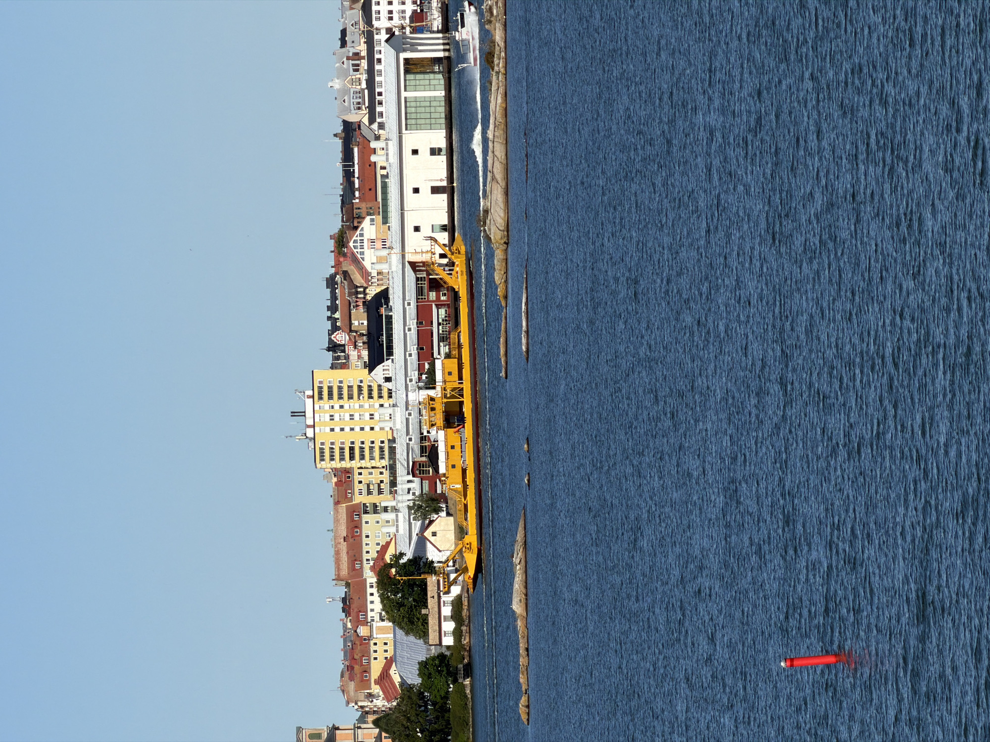 The image depicts a sunny day in Karlskrona, Sweden, with a clear blue sky above. In the foreground, the deep blue water of the sea is visible, with small waves rippling across its surface. A bright red buoy with a white top stands out in the water on the left side of the frame. In the middle ground, a long, yellow ferry is navigating through the water, leaving a white wake behind it. To the right of the ferry, a smaller white boat is moving at speed, also creating a wake. Scattered in the water are several rocky islands, some with sparse vegetation.
The background reveals the city skyline of Karlskrona. A row of buildings, varying in height and architectural style, lines the waterfront. Some structures are taller, modern apartment buildings, while others appear to be older, with red-tiled roofs. Several cranes are visible, suggesting ongoing construction or industrial activity. The overall impression is of a bustling port city on a pleasant day.