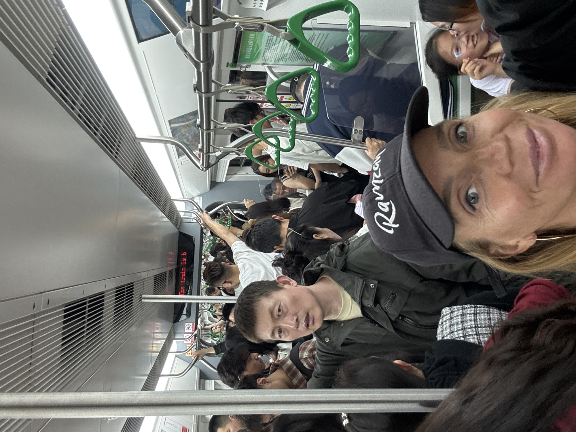 The image depicts a crowded subway car, likely in the city of Yuzhong District, China. Passengers are tightly packed together, some holding onto green-handled bars suspended from the ceiling. A woman in a dark baseball cap is taking a selfie. Another woman with glasses is standing nearby. Many other individuals are visible, indicating a high volume of commuters. The interior of the subway car is modern, with white walls and metallic fixtures. The subway car shows "The train is b".