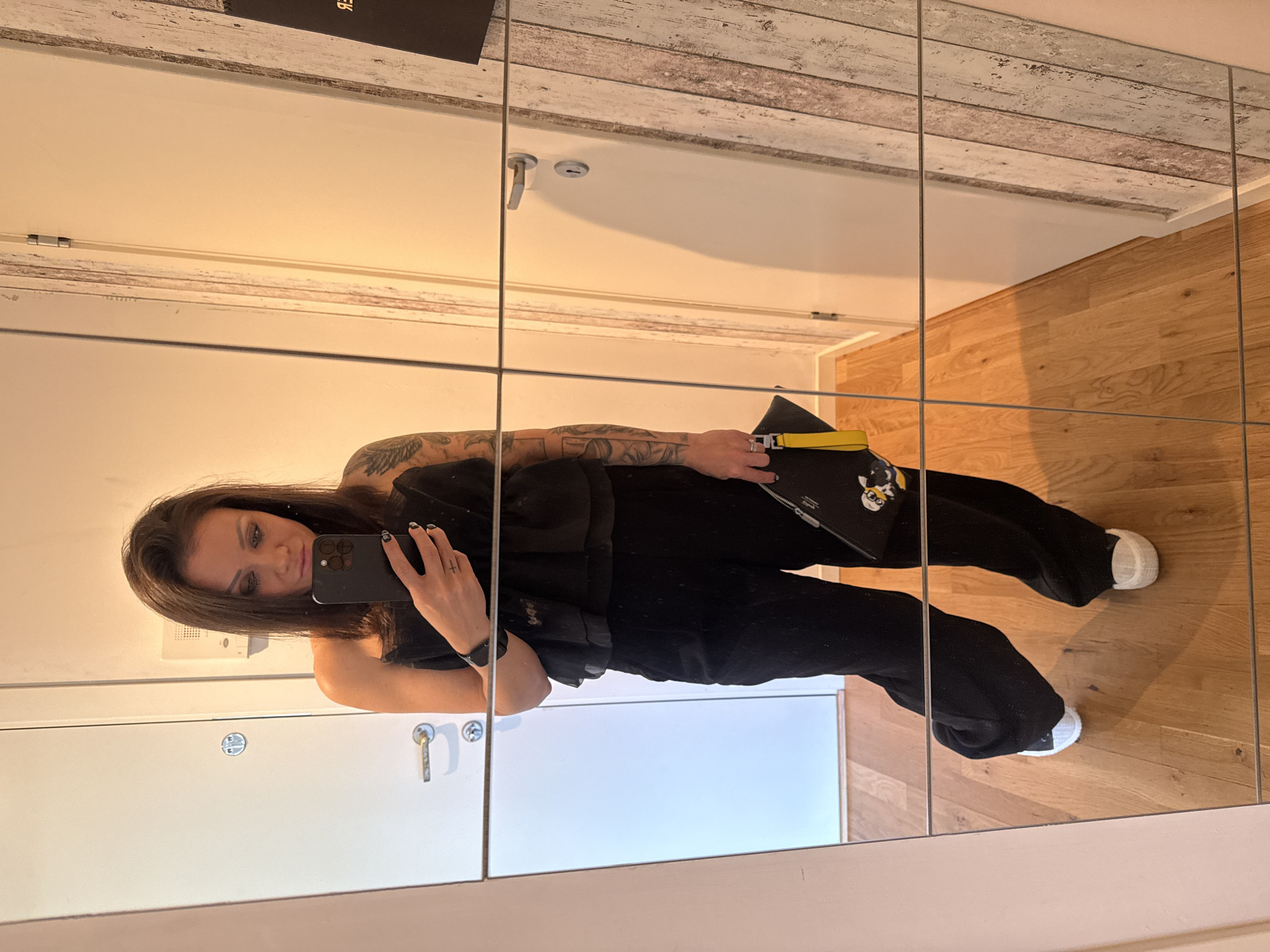 A person is taking a picture of themselves in a mirror. The person is wearing a black outfit and white shoes, and is holding a black phone in their right hand. They also hold a black purse with a yellow strap, decorated with what looks like an anime character. The location appears to be a room with wood floors and white walls. The room is inside a building somewhere in the city of Unknown, Unknown.