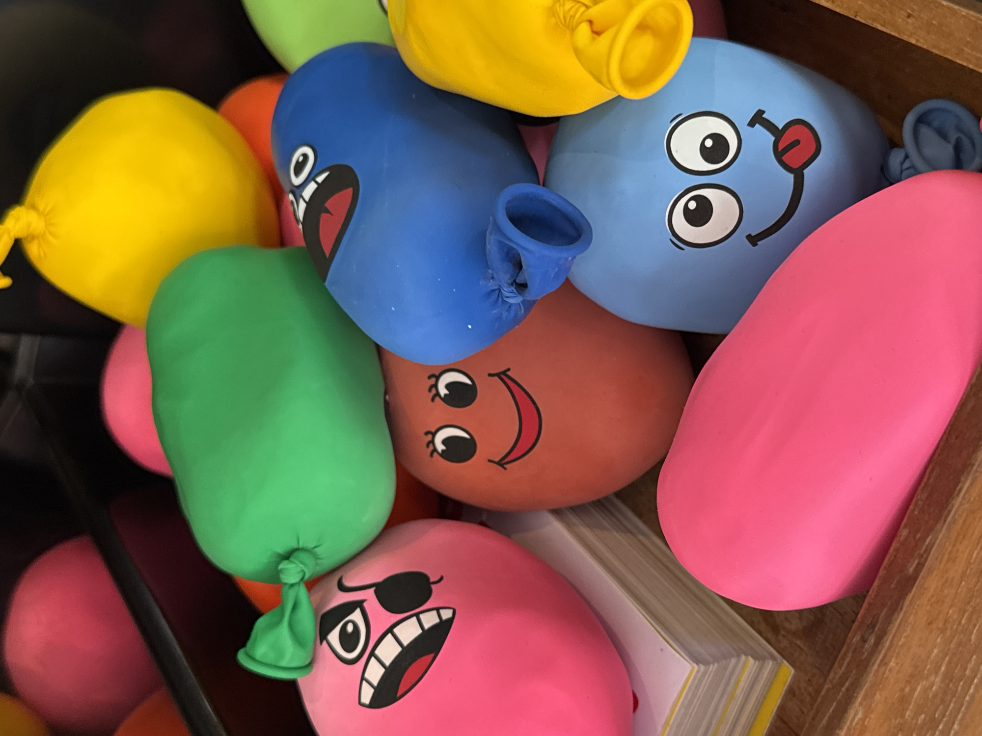A high-angle, close-up shot displays a collection of inflated latex balloons with printed cartoon faces, contained within a wooden box. The balloons are in various colors, including yellow, blue, green, red, and pink. Each balloon features a different facial expression, ranging from happy and silly to angry and pirate-like. A stack of books sits partially obscured beneath some of the balloons. The location is not discernible from the image.
