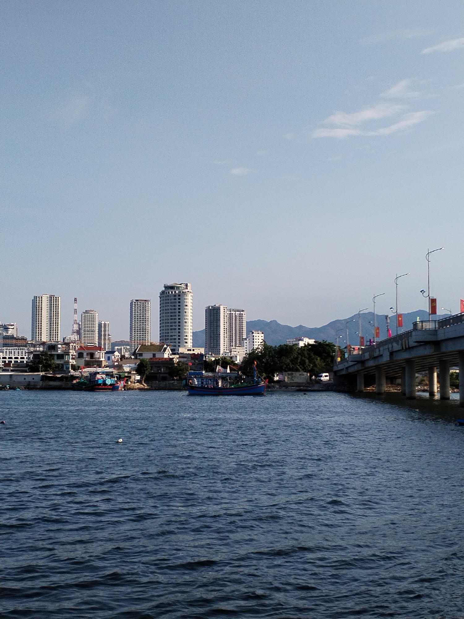 The image presents a waterfront scene with the skyline of a city, likely in or near Bac Nha Trang Ward, Vietnam, given the flags on the bridge. The foreground is dominated by dark blue water with visible ripples and movement. A bridge, adorned with streetlights and red flags, spans the water on the right side of the frame. Buildings, including several tall skyscrapers, form the cityscape in the middle ground. In between the water and buildings, there are smaller structures, trees, and at least two boats. The horizon is marked by the outline of hills or mountains in the background. The sky is pale blue with a few faint clouds.