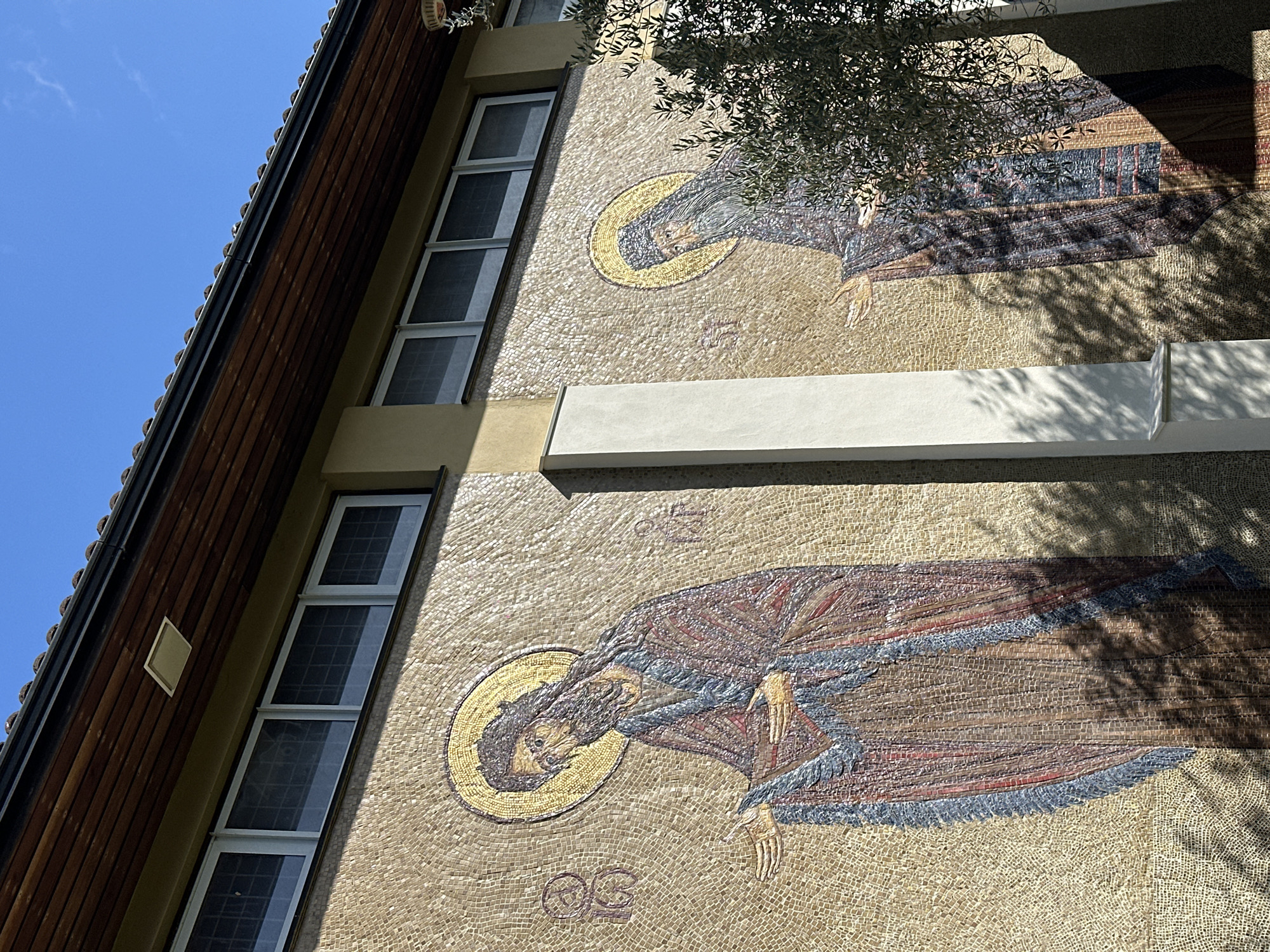 The image displays the exterior south-facing mosaic-adorned wall of The Holy Fathers of Nicaea Orthodox Church, located on London Road, Maldon, United Kingdom. The wall features two large, vertical mosaic icons against a background of light beige tesserae. On the left, a bearded male figure, depicted as a saint with a golden halo and dark hair, is shown wearing a red and blue robe with open hands, extending to the right. Inscriptions, consistent with "IC XC," are visible to his left. A vertical white architectural pillar separates this mosaic from the right section. On the right, a second bearded male figure, also a saint with a golden halo, is depicted in a dark robe, partially obscured by the dark green foliage of an olive tree. His hands are also extended, mirroring the left figure. Above the mosaics, two distinct sections each contain four horizontal rectangular windows with white frames and dark grilles. Above the windows, a light beige wall section transitions to a dark brown wooden soffit with horizontal planking, which incorporates a white rectangular vent on the left. A black gutter runs beneath the red-brown tiled roofline. Strong sunlight illuminates the scene, casting distinct shadows from the olive tree onto the mosaics and the architectural pillar against a clear blue sky.