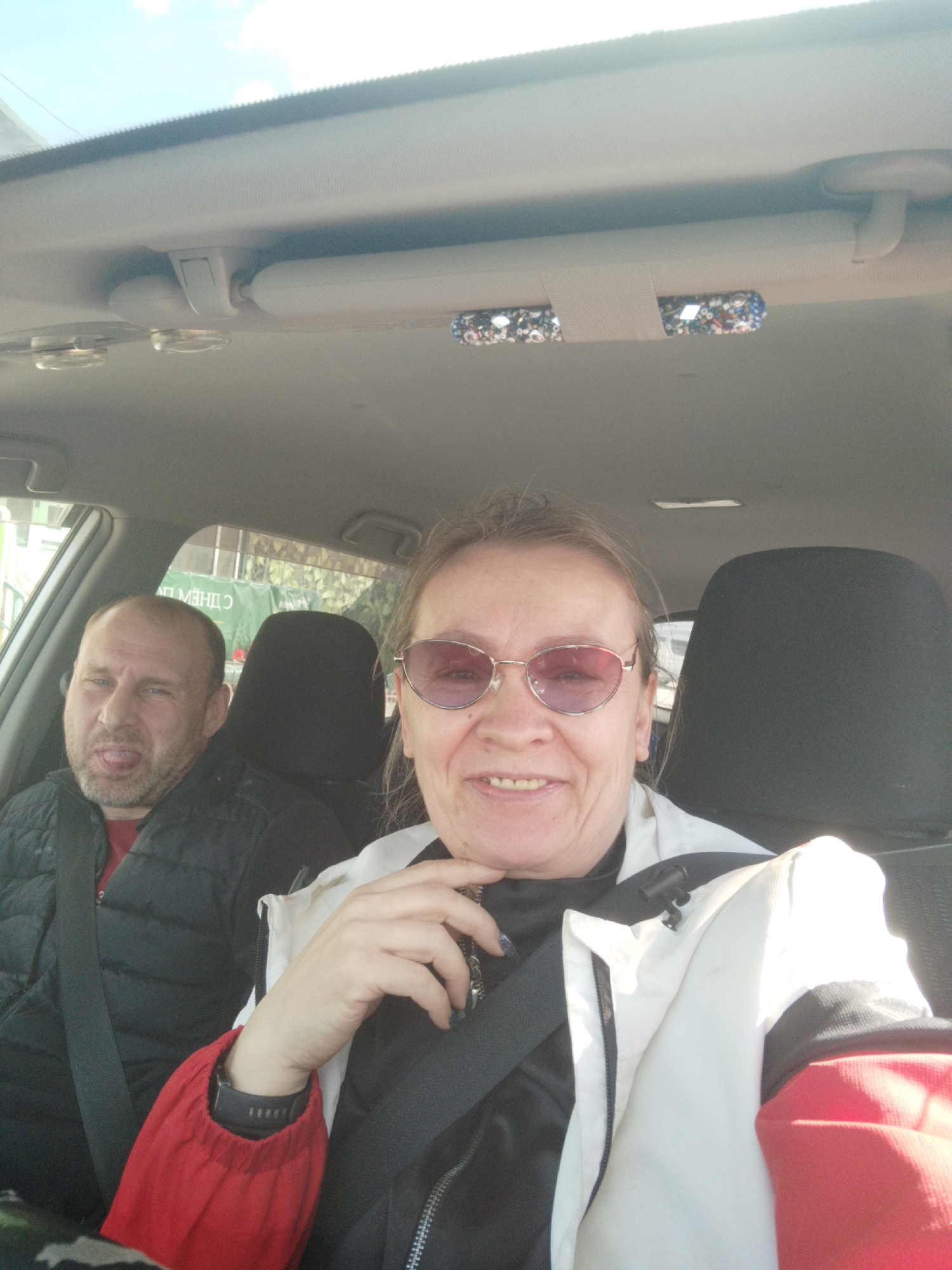 Two people are in a car, possibly in Yakutsk, Russia. The woman in the foreground is wearing tinted glasses, a black top, and a red, white, and black jacket. She is smiling slightly and has her hand near her chin. She is wearing a black smartwatch. The man in the background is sticking his tongue out and wearing a black puffer jacket over a red shirt. Both are wearing seatbelts. The car's interior is light gray. There are two lights on the car's ceiling. A green sign with white Cyrillic lettering is visible outside the window on the left.