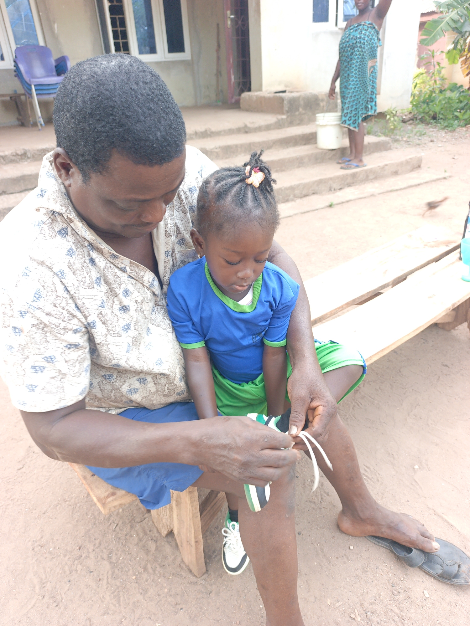 The image portrays an outdoor scene in Akwanga, Nigeria, during the daytime. A man and a young girl are seated on a wooden bench, with the man actively helping the girl with her shoe laces. He wears a patterned short-sleeved shirt and blue shorts, while the girl is dressed in a blue top with green trim and green shorts, along with white and green sneakers. Both individuals are looking down, focused on the task at hand.
In the background, a building with white windows and steps is visible. A woman in a patterned blue and green dress or wrap stands on the steps, holding a white bucket and raising one arm. Nearby, a stack of blue plastic chairs is seen. The ground is bare earth, and green foliage, including what appears to be a banana tree, contributes to the natural setting. A small bird is faintly visible on the ground. The scene conveys a calm, everyday moment under clear, sunny weather conditions. No visible text is present in the image.