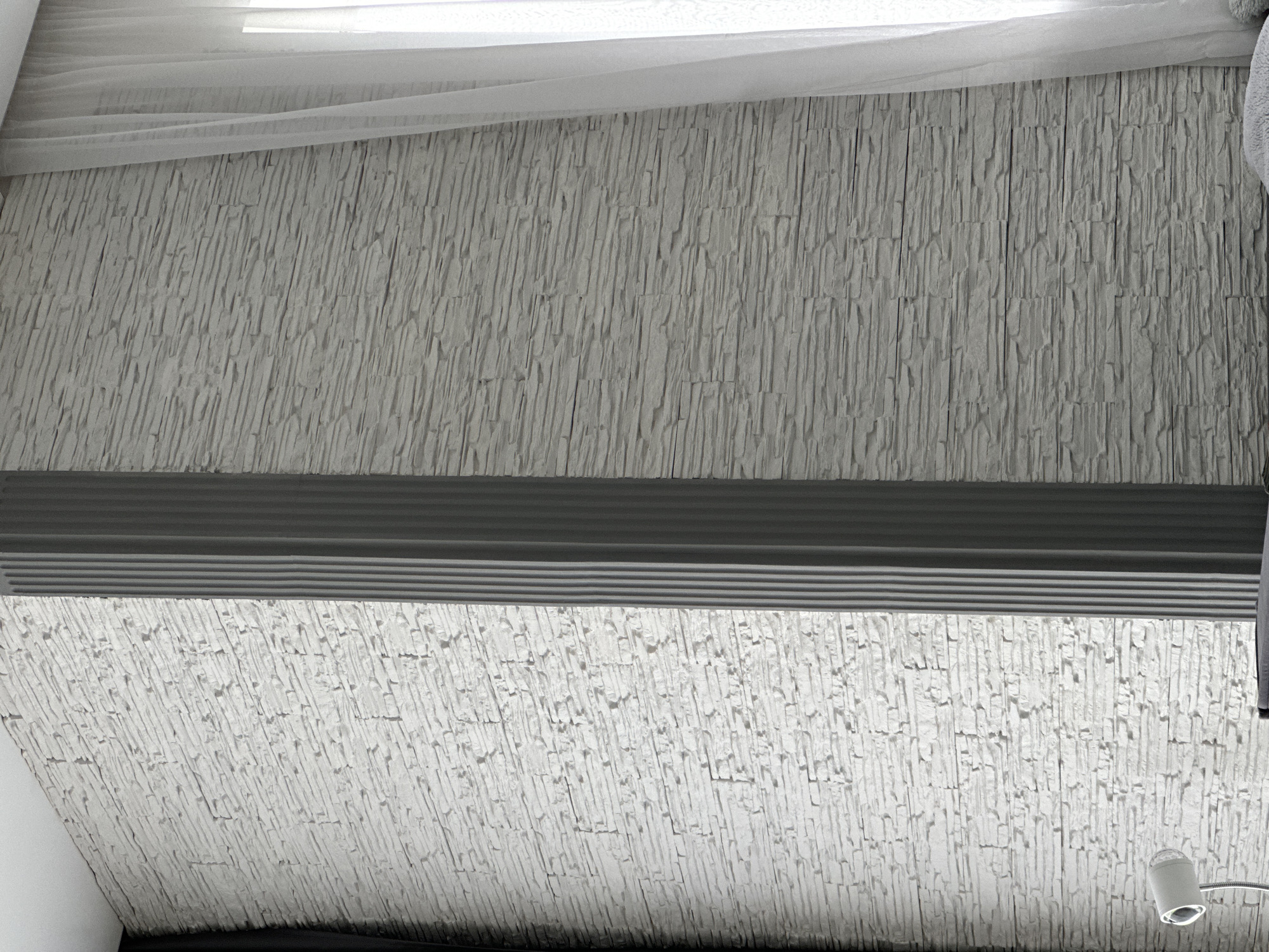 The image displays an interior architectural view, primarily focusing on a textured wall or ceiling. The dominant surface is covered with irregular, elongated, and vertically aligned light grey or white elements, resembling stacked stone veneer or a textured paneling.
A prominent dark grey horizontal band with several subtle, parallel ridges runs across the middle of the frame. This band could be a vent, a window blind, or a decorative architectural feature.
Above this dark band, the textured surface continues upwards, partially obscured by flowing, sheer white fabric, likely curtains or drapes. Bright light emanates from behind this fabric, suggesting the presence of a window or skylight, indicating daytime.
Below the dark horizontal band, the same textured surface is visible again, but it appears significantly brighter and more illuminated than the upper section, possibly due to direct light. In the bottom right corner, a white lamp fixture with a spherical, possibly faceted, head is visible, attached to a flexible silver arm. A small portion of a plain white wall is also visible in the bottom left corner.
The scene is static, with no visible people or activities. There is no discernible text within the image. The overall impression is of a modern, brightly lit indoor space, possibly featuring a sloped ceiling or a high wall.