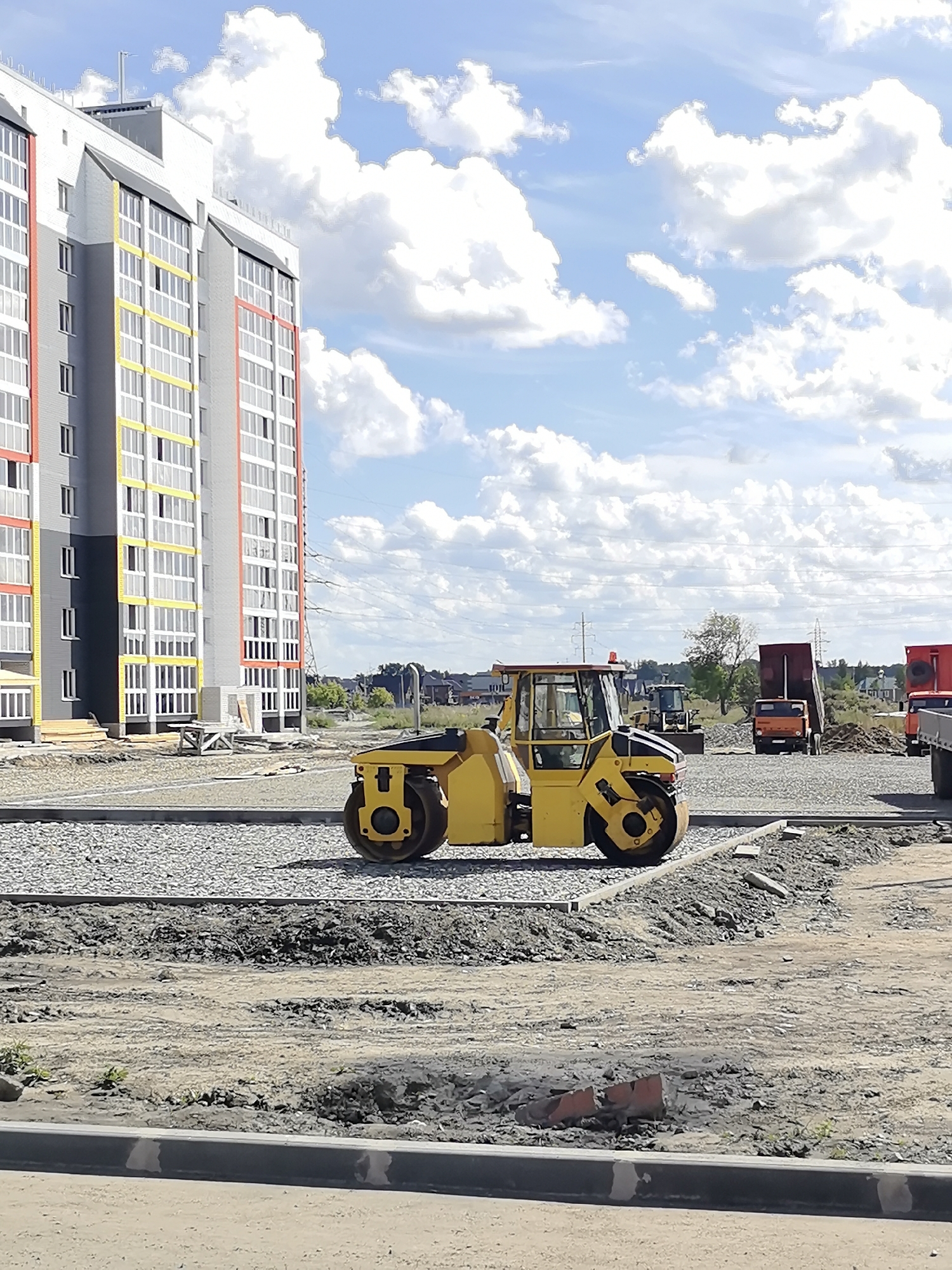 The image depicts a construction site in Novoaltaysk, Russia. In the foreground, a bright yellow steamroller is parked on a gravel surface. Behind it, a tall, modern apartment building with white, gray, yellow, and red accents is under construction. The sky is blue with scattered white clouds. Further in the background, various construction vehicles, including trucks and a bulldozer, are visible, along with trees and smaller buildings, suggesting a developing urban area. The ground is a mixture of gravel, dirt, and what appears to be partially laid asphalt. There are no people visible in the image.