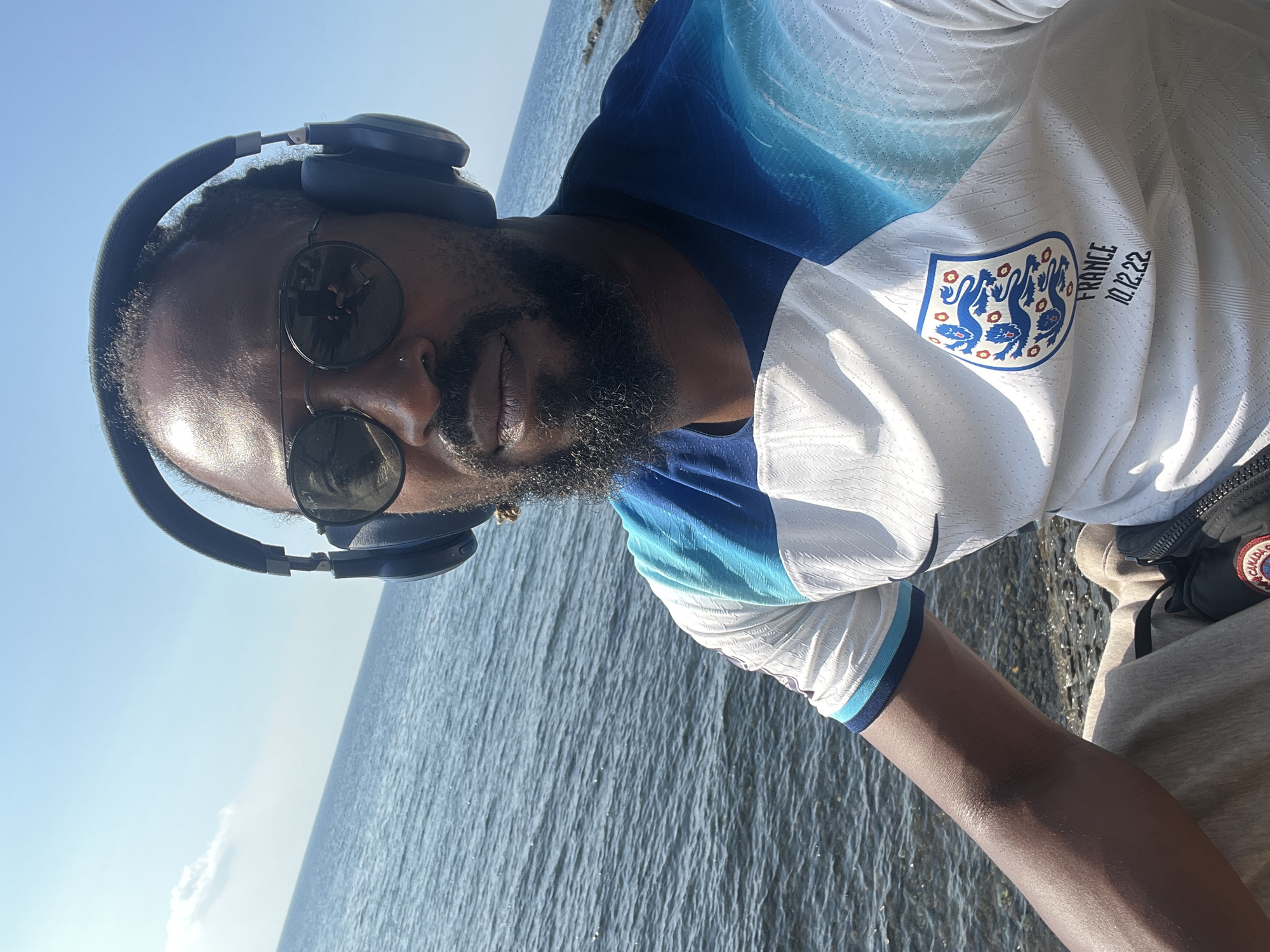 The media file is a close-up, selfie-style shot of a man wearing headphones and sunglasses, facing the camera. He has a beard and is wearing a white and blue patterned t-shirt with a crest and text that reads "FRANCE 10.12.22". The background is a body of water under a clear blue sky, suggesting an outdoor setting. The lighting indicates it is daytime. The man's expression appears neutral. The presence of the t-shirt with a date might suggest a connection to a specific event or team. The water appears calm, with some texture from ripples. The overall impression is of a casual, possibly contemplative moment outdoors.
