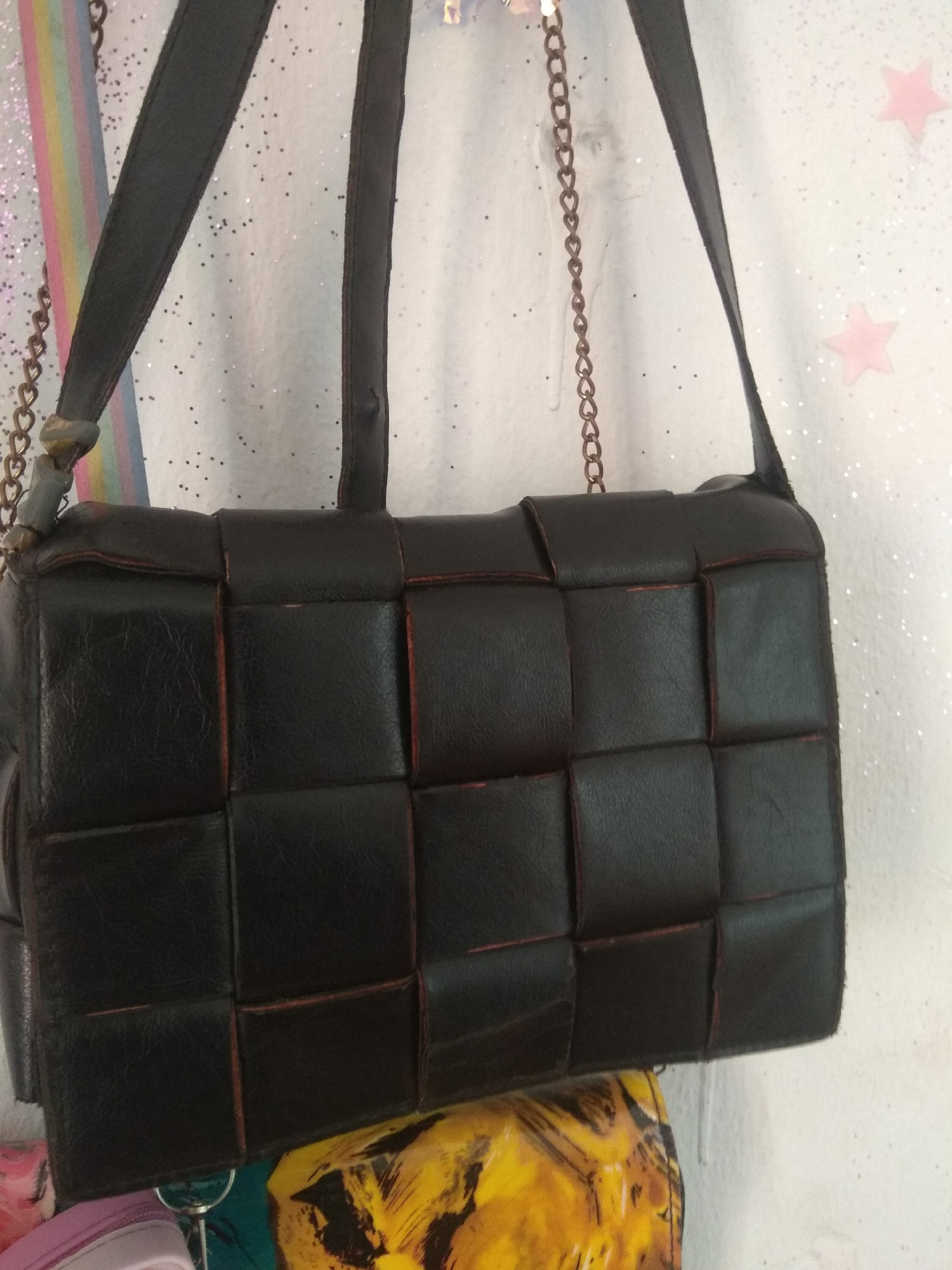 The media file displays a close-up of a dark, woven leather handbag. The handbag has a structured, rectangular shape with a woven pattern formed by alternating strips of leather. The edges of the leather strips reveal a reddish-brown undertone. The handbag is suspended by dark straps, one of which is a thin leather strap, and another appears to be a chain with heart-shaped links. The background is a white wall adorned with scattered, subtle glitter and two faint pink star shapes. To the left of the handbag, a portion of a colorful, rainbow-striped fabric is visible. Below the handbag, there are hints of other items, including what appears to be a portion of a colorful garment or bag with yellow and black patterns, and the edge of a pink item. The lighting suggests an indoor setting, likely during the daytime. There is no discernible text in the image. The overall presentation is that of a product shot or a casual display of a fashion accessory.