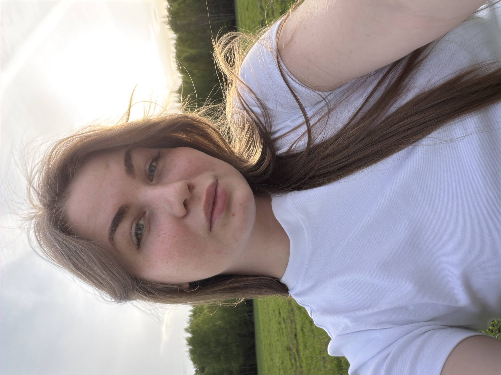 A young woman with light brown hair and freckles is taking a selfie outdoors. She is wearing a white t-shirt. The background shows a grassy field and trees, suggesting a park or rural area. The lighting suggests it may be late afternoon or early evening. The specific location within the city of Zlodar', Russia cannot be determined from the image.