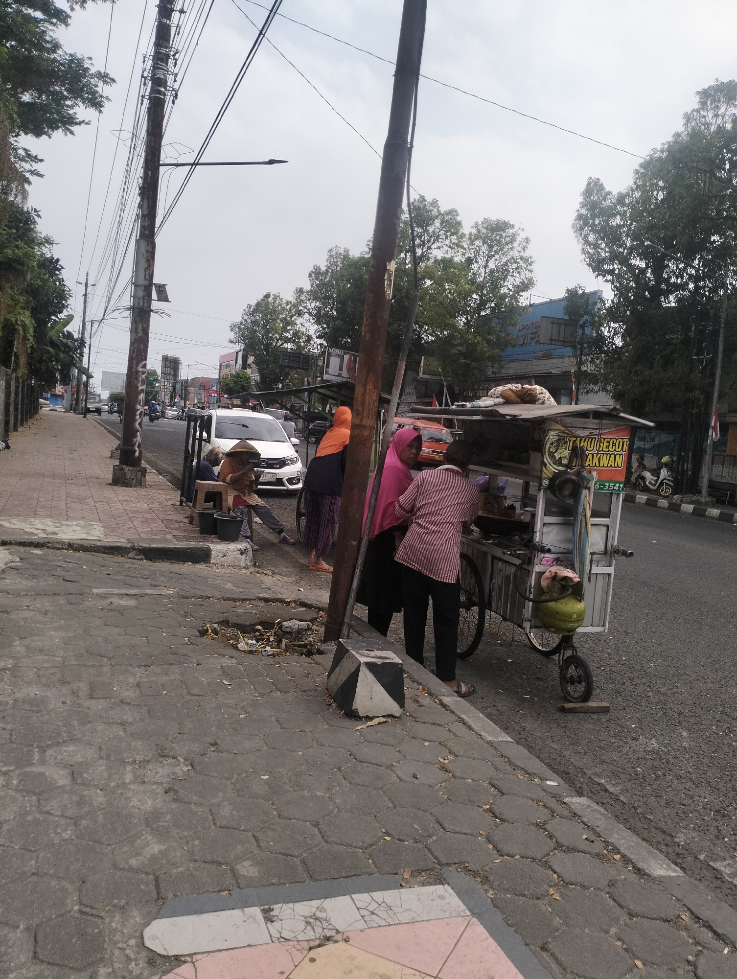 The image shows a street scene in Purbalingga, Indonesia. On the right side of the image, a food cart is parked along the roadside. The cart has a white and silver frame with a blue awning. There are people around the cart, some appear to be customers or vendors. One person is wearing a pink headscarf and a striped shirt, and another is wearing a light-colored headscarf. A gas cylinder is attached to the front of the cart. In the background, there is traffic on the road, including cars and motorcycles. A white car is prominently visible in the middle ground. Power lines and utility poles run along the side of the street. On the left side of the image, a sidewalk is visible, paved with hexagonal and rectangular bricks. There are also trees and buildings in the background. A person wearing a conical hat is sitting on a stool near the food cart. The overall scene depicts a typical street vending operation in an urban environment.