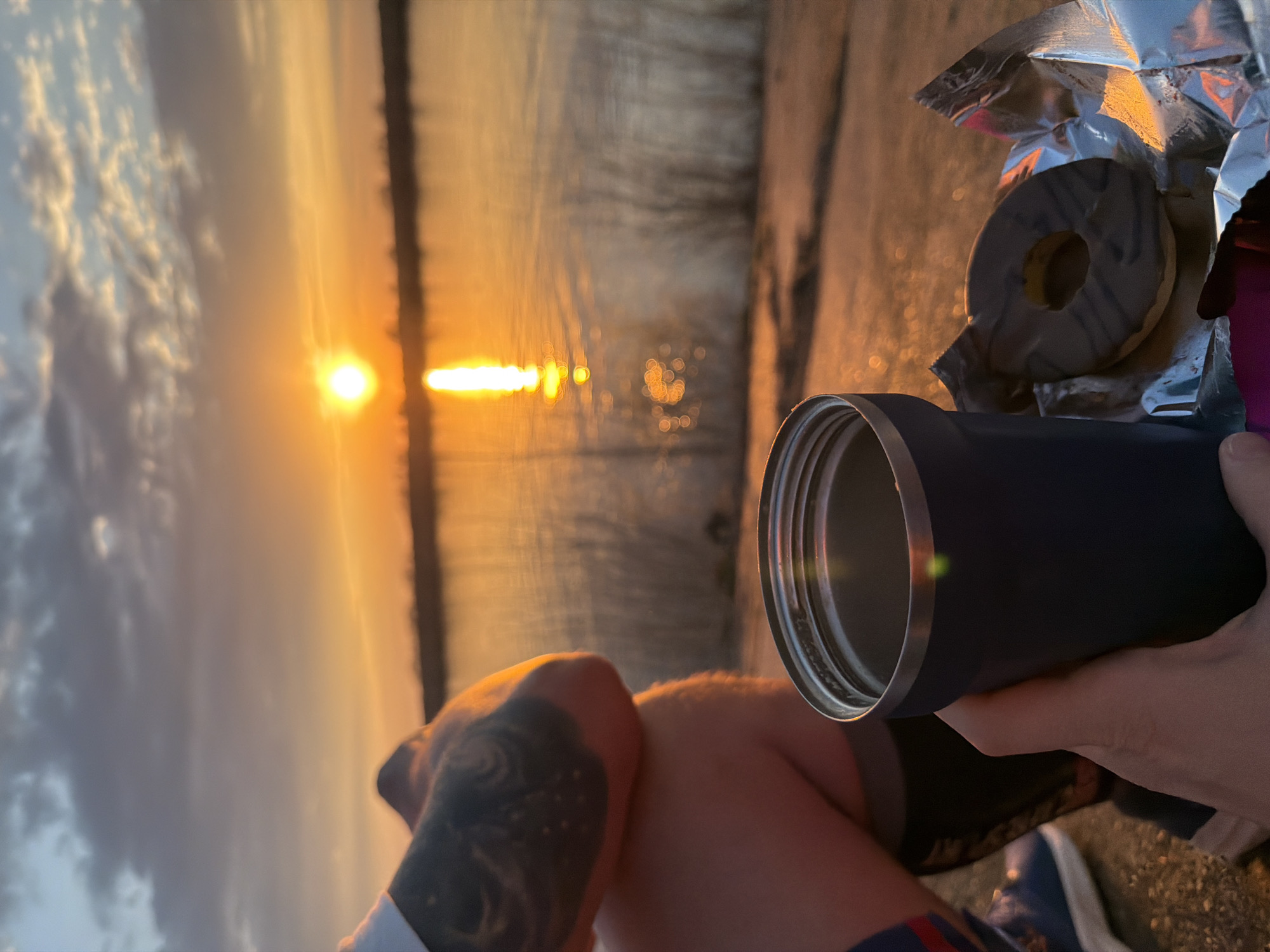A person sits near a body of water at sunset. The location is Chaikovsky, Russia. The sun is setting over the water. The person is holding a dark blue tumbler in their right hand and resting their legs, one with a tattoo, on top of each other. Next to the tumbler is an opened package of Oreo cookies. The person is wearing shorts and blue athletic shoes.