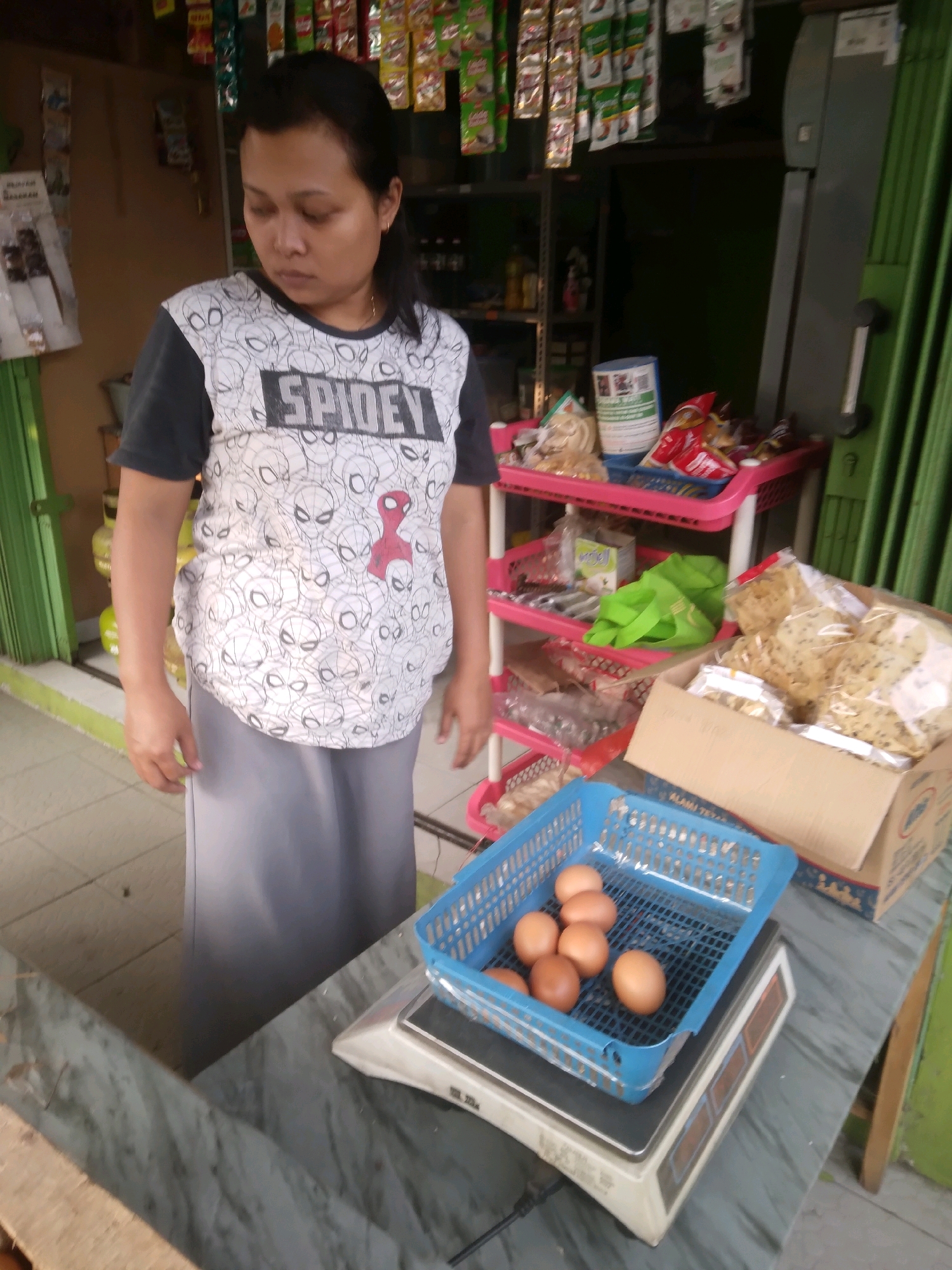 A woman stands in front of a small shop in Desa Pamijen, Kecamatan Sokaraja, Indonesia, looking down at a digital scale. The scale holds a blue plastic basket containing seven brown eggs. The woman wears a t-shirt with a Spider-Man pattern and a gray skirt. Behind her, shelves are stocked with various packaged food items and bags, including what appears to be crackers in a cardboard box. The shop has a green metal shutter on the right side. The floor is tiled and a yellow propane tank is visible to the left.