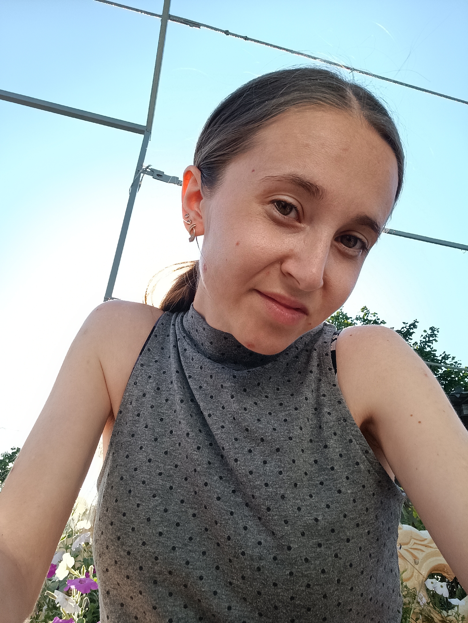 A woman is taking a selfie outdoors, under a clear blue sky. She is wearing a grey sleeveless top with a polka dot pattern and a high neckline. She has a small, decorative earring in her left ear. In the background, there is a metal structure, some greenery, and what appears to be flowers. The location is Vypasne, Ukraine.
