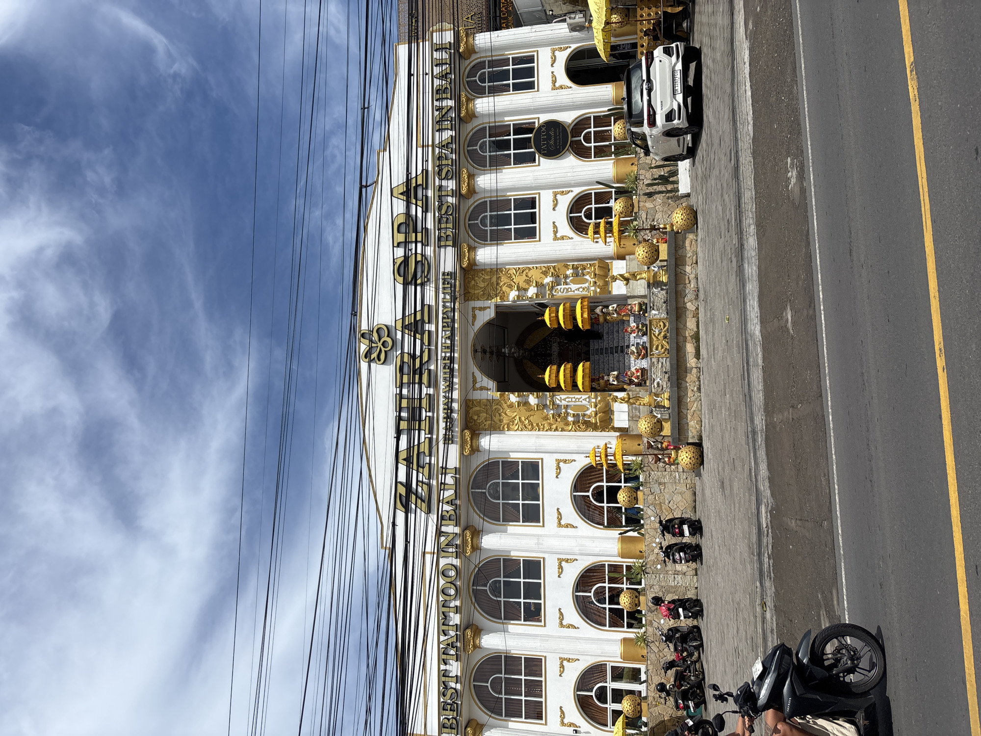 The image displays a bustling street scene in front of a highly ornate commercial building. The main subject is a large, multi-story building, predominantly white with extensive gold decorative accents, including columns, archways, and sculpted details. Overhead, numerous black power lines or communication cables crisscross against a bright blue sky with wispy white clouds.
The building's facade prominently features signs indicating its businesses. Visible text includes "BEST TATTOO IN BALI" and below it, "THE HAPPY LIFE". Another large sign, partially obscured by wires, reads "ARTAWAN BEST SPA IN BALI". A circular sign on the building's exterior also clearly states "TATTOO Studio". The entrance area is adorned with golden decorative spheres and yellow traditional Balinese-style umbrellas.
In front of the building, a paved sidewalk leads to a road with a distinct yellow dividing line. Several vehicles are parked along the sidewalk and street. A white car with the license plate "DK 1267 FCO" is parked close to the building. Multiple motorcycles and scooters, including one with "HONDA" visible on its side, are parked along the sidewalk, suggesting a common mode of transport in the area. A person is partially visible on a motorcycle in the foreground.
The scene takes place during the daytime under clear and fair weather conditions. The architectural style, the presence of multiple scooters, and the explicit mention of "BALI" in the signage strongly suggest the location is in Bali, Indonesia, aligning with the provided context of Kuta Selatan. The overall impression is that of a lively tourist or commercial area.