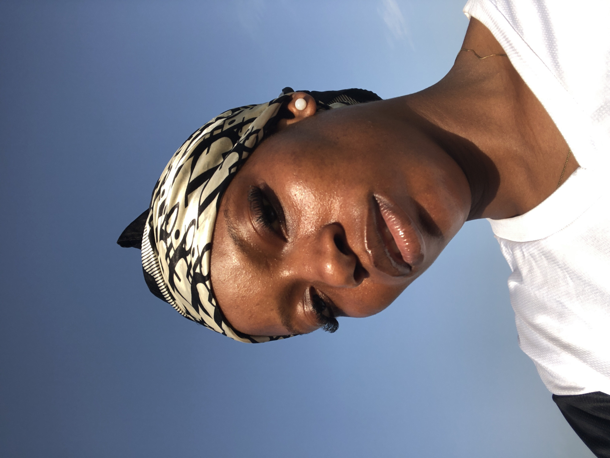 The image is a close-up, low-angle shot of a young Black person's face, tilted upwards against a clear blue sky. They are wearing a black and white patterned headscarf that covers their hair and a white t-shirt. A pearl earring is visible in their left ear. The lighting suggests it is daytime, with the sun casting a warm glow on their skin and highlighting their glossy lips and prominent eyelashes. Their eyes are mostly closed, and their expression appears serene or contemplative. A thin gold necklace is visible on their neck. The background is a solid, cloudless blue sky, giving a sense of being outdoors and possibly looking up from a lower vantage point. The orientation of the image is rotated 90 degrees counter-clockwise.