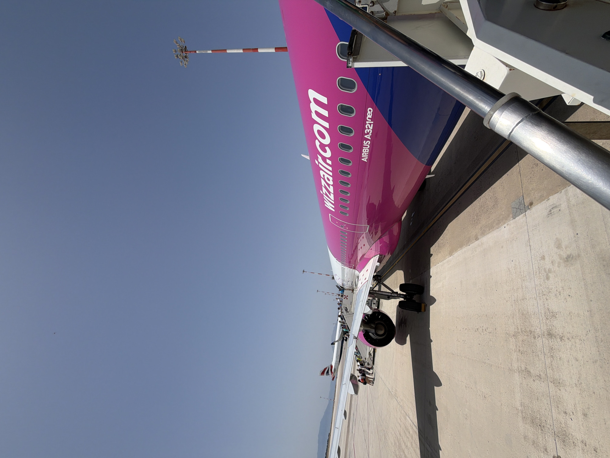 This is a close-up, low-angle, eye-level outdoor shot of a Wizz Air Airbus A321neo airplane at the Palermo Airport in Cinisi, Italy. The plane is predominantly bright pink with a dark blue stripe along the bottom of the fuselage. The Wizz Air logo and website, "wizzair.com," are clearly visible on the side of the plane in white text. Below the windows, the text "AIRBUS A321neo" is also visible.
The foreground features a silver metal railing and a section of white stairs, suggesting a mobile boarding stair or jet bridge. The plane's landing gear is visible, resting on a concrete tarmac. In the background, another airplane, identified as a British Airways aircraft, is parked with passengers boarding via stairs. Several people are seen disembarking or boarding the Wizz Air plane.
The environment is an airport tarmac on a clear, sunny day. The sky is a vibrant, cloudless blue. A red and white striped airport light pole is visible in the upper right corner of the frame. The overall impression is a busy airport scene during daylight hours.