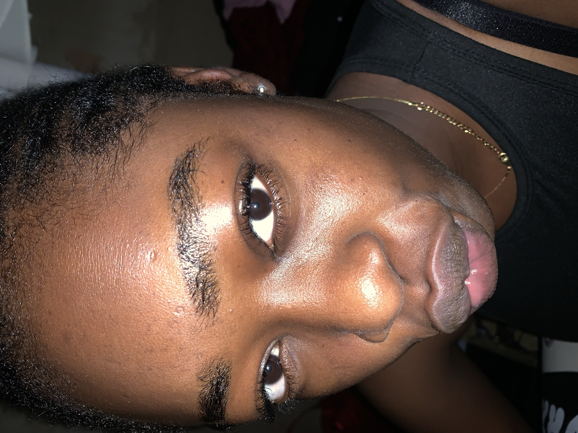 The image is a close-up portrait of a young Black woman's face, taken indoors. Her skin is dark brown and appears slightly dewy, with visible pores and a few small blemishes on her forehead and cheeks. She has dark, natural hair styled close to her scalp, with some wisps around her hairline. Her eyebrows are dark and well-defined. Her eyes are dark brown and wide open, with long, dark eyelashes. Her nose is broad and well-defined. Her lips are full and slightly parted, with a rosy tint. She is wearing a gold chain with small pendant-like details around her neck and a black sleeveless top. The lighting suggests an artificial light source, casting some highlights on her face. The background is dark and out of focus, with hints of fabric textures and possibly furniture. There is no indication of the location being in the city of Rafin Par, Nigeria.