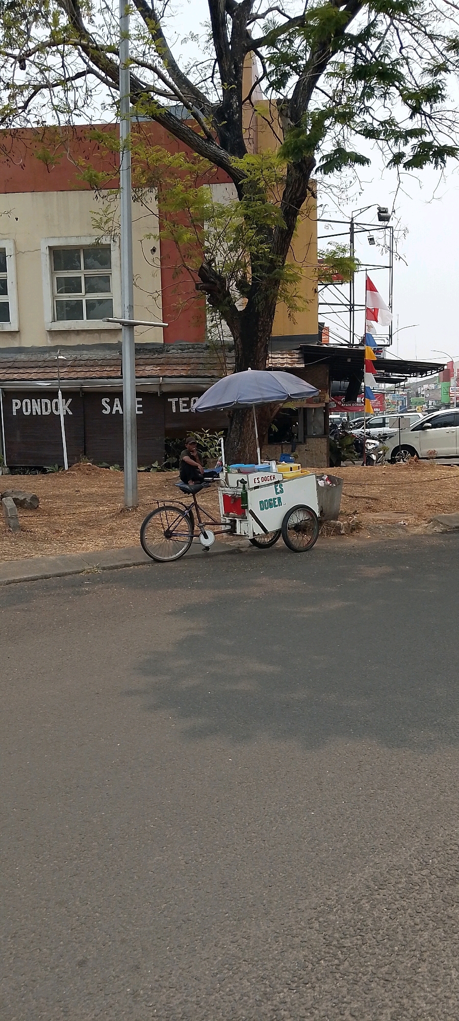 A tricycle ice cream cart is parked on the side of a road in Cileungsi, Indonesia, under a large tree with sparse green leaves. The cart is white with green lettering that reads "ES DOGER" and has a blue umbrella providing shade. A man wearing a dark t-shirt and dark pants is seated on the cart's saddle, appearing to be smoking and looking to his left. The cart has two rear wheels and one front wheel, with various containers and items visible on its top surface. Behind the cart, a building with orange and cream colored walls and windows is partially visible. The ground next to the road is covered in dry grass and dirt. In the background, there are flags, traffic lights, and other vehicles, including a white car and what appears to be a motorcycle. The sky is overcast.