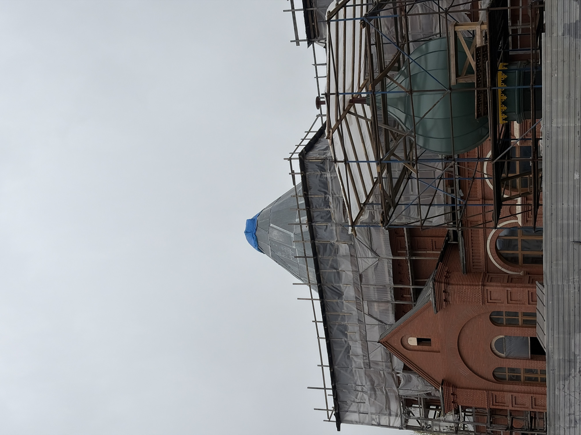 The image captures a historic-looking building, likely in Saint Petersburg, Russia, undergoing extensive renovation or construction. The building is constructed of warm red brick, featuring intricate architectural details, including multiple arched windows with visible wooden frames and decorative brickwork along its walls.
A prominent feature is a large dome or spire, partially visible through scaffolding and protective coverings, which appears to be a distinct green or teal color. Extensive scaffolding, made from a combination of metal and wooden poles and frames, encases much of the building's exterior. Sections of the scaffolding and the building are draped with translucent plastic sheeting and dark tarpaulins, presumably for weather protection or dust containment. A bright blue tarp covers the top of one conical roof section, suggesting recent work or a focus point of the renovation.
The sky overhead is uniformly overcast and light grey, indicating a cloudy day. No people are visible in the scene, and there is no discernible text. The comprehensive scaffolding and protective coverings clearly signify that construction, restoration, or maintenance work is actively taking place on the structure.