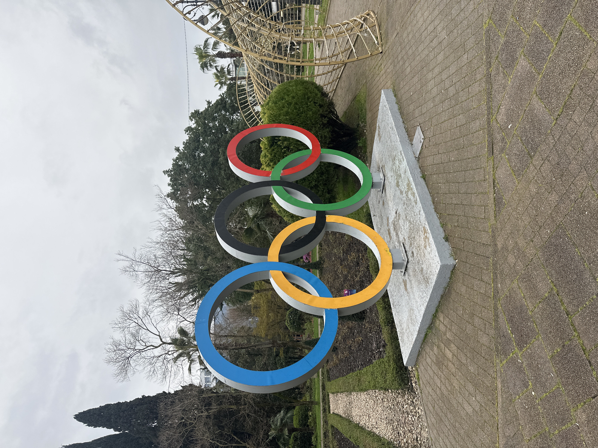 The image captures a statue of the Olympic rings in Sochi, Russia. The rings, colored blue, black, red, yellow, and green, are mounted on a concrete base. The background includes trees, a wrought-iron structure, and a brick-paved area. The sky is overcast. There are no people visible in the frame.