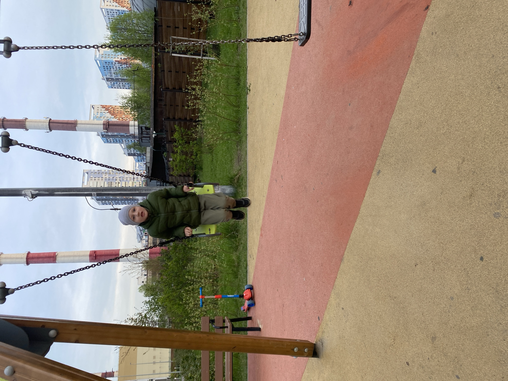 The photograph depicts a young child sitting on a swing in a playground. The child is wearing a green jacket, khaki pants, and a gray hat. The playground has a red and yellow surface. A scooter is visible on the ground near the swing. In the background, there are tall industrial chimneys painted with red and white stripes, along with buildings typical of Saint Petersburg, Russia. The overall impression is of a playground located in an urban area.