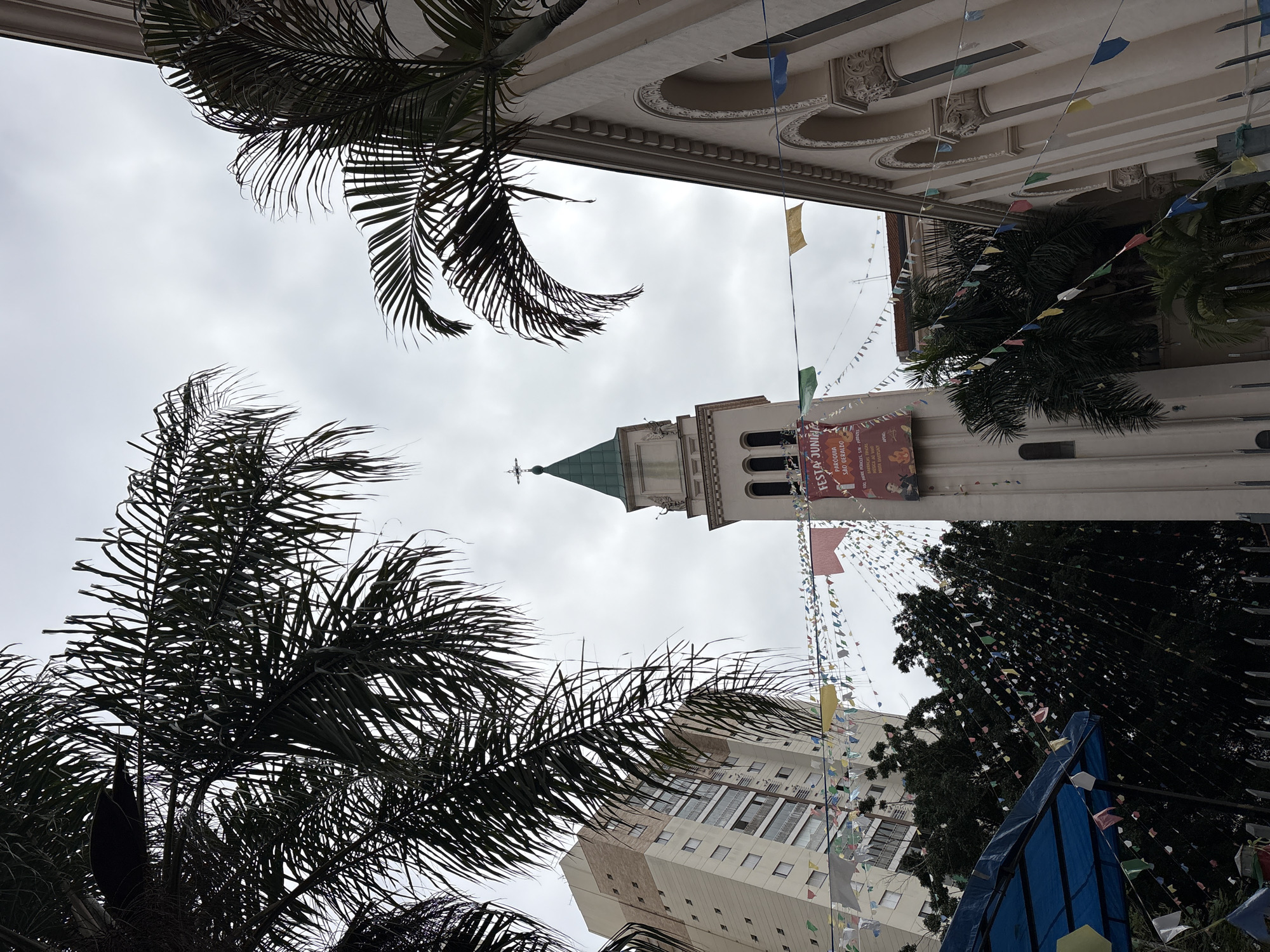 The image captures a street view in São Paulo, Brazil, likely during a festival. In the foreground, palm fronds frame the scene, with the fronds of two large palm trees partially obscuring the view of buildings and a church tower.
Dominating the mid-ground is a church tower with a green, pointed roof, adorned with a cross at its apex. A banner hangs from the tower, displaying text that includes "FESTA JUNINA" and "PARÓQUIA SÃO GERALDO", indicating a June Festival at the São Geraldo Parish. The church facade to the right features arched windows and decorative architectural elements.
In the background, a modern high-rise building with multiple windows stands tall. Strung between the buildings and the church are numerous colorful triangular flags, typical of Brazilian June Festivals, and some larger rectangular banners. Below the church tower, on the left side of the image, a blue awning is visible, suggesting a temporary stall or shelter. The sky is overcast, contributing to the diffused lighting. No people are distinctly visible in the scene. The overall atmosphere suggests a festive urban environment.